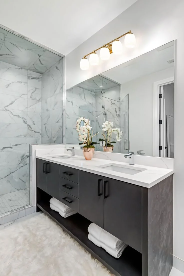 Modern bathroom with a double vanity, large mirror, marble walls in the shower, and white towels on open shelves.