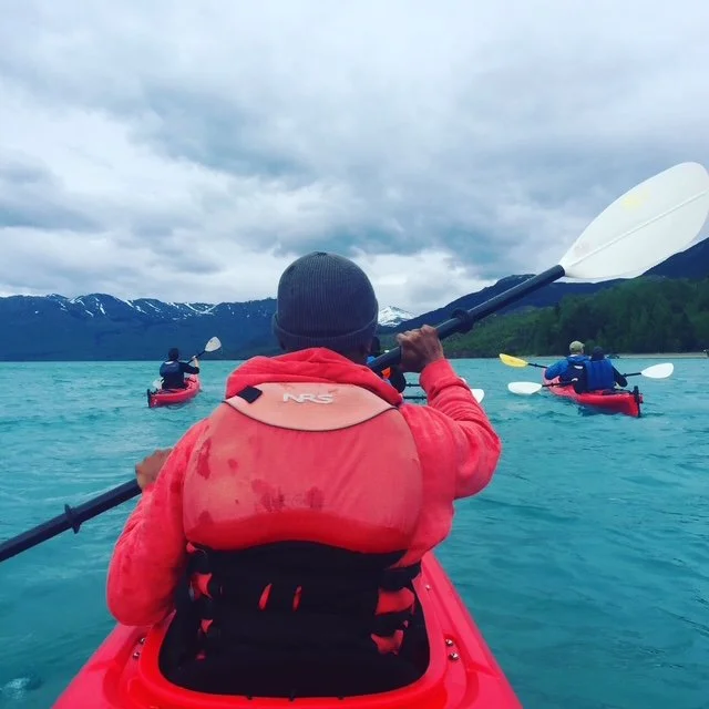 A person in a red kayak with a life jacket paddling on a lake with other kayakers, surrounded by mountains and cloudy sky.