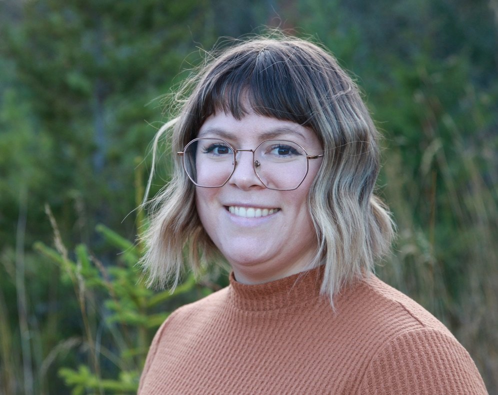 A woman with shoulder-length wavy hair, wearing glasses and an orange sweater, smiling outdoors with green foliage in the background.