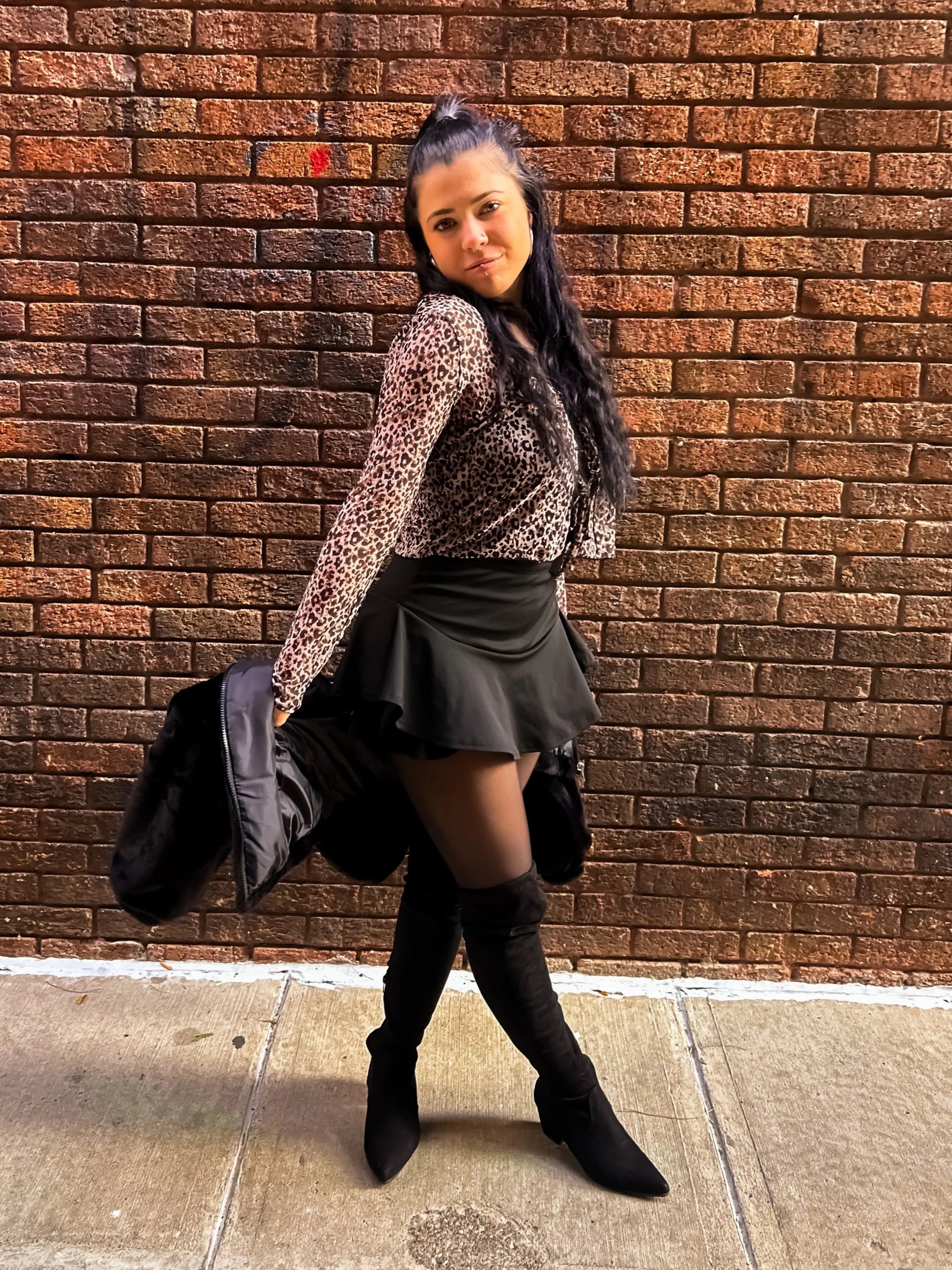 Young woman posing against a brick wall, holding a black jacket, wearing a leopard print top, black skirt, black tights, and over-the-knee black boots.