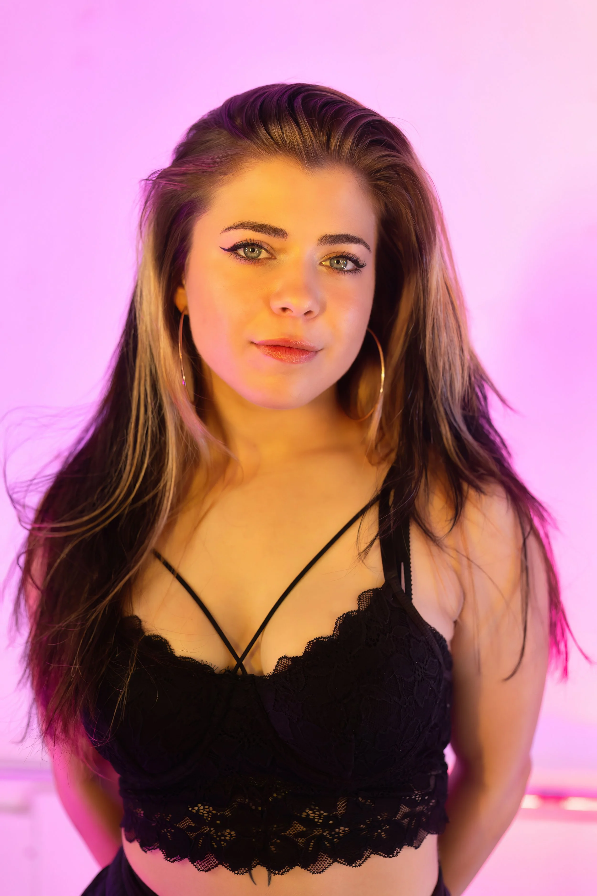 A young woman with long, wavy brown hair and green eyes, wearing hoop earrings and a black lace crop top, standing against a pink background.