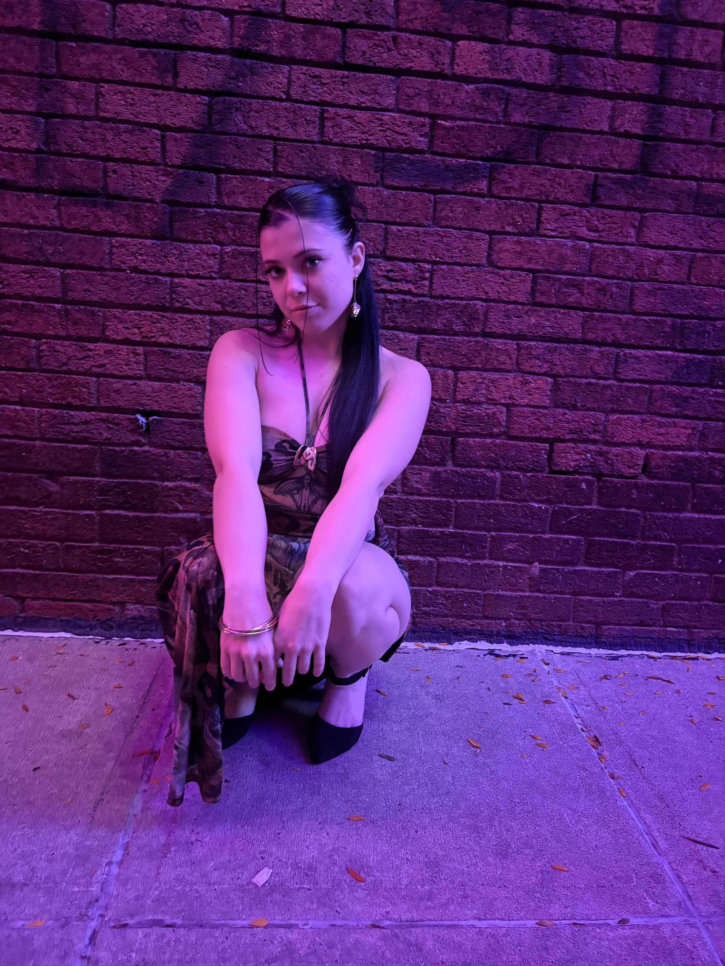 Young woman with long dark hair in a dress squatting against a brick wall illuminated by purple lighting.