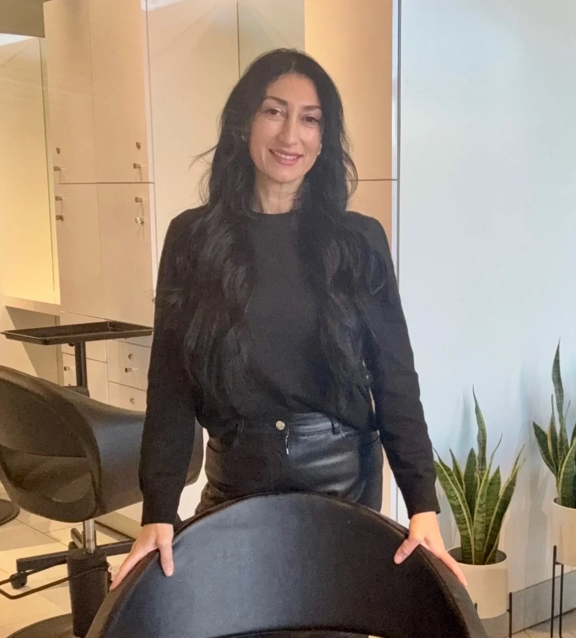 Woman with long black hair standing in an office or meeting room, wearing a black top and leather pants, smiling at the camera with office furniture and houseplants in the background.