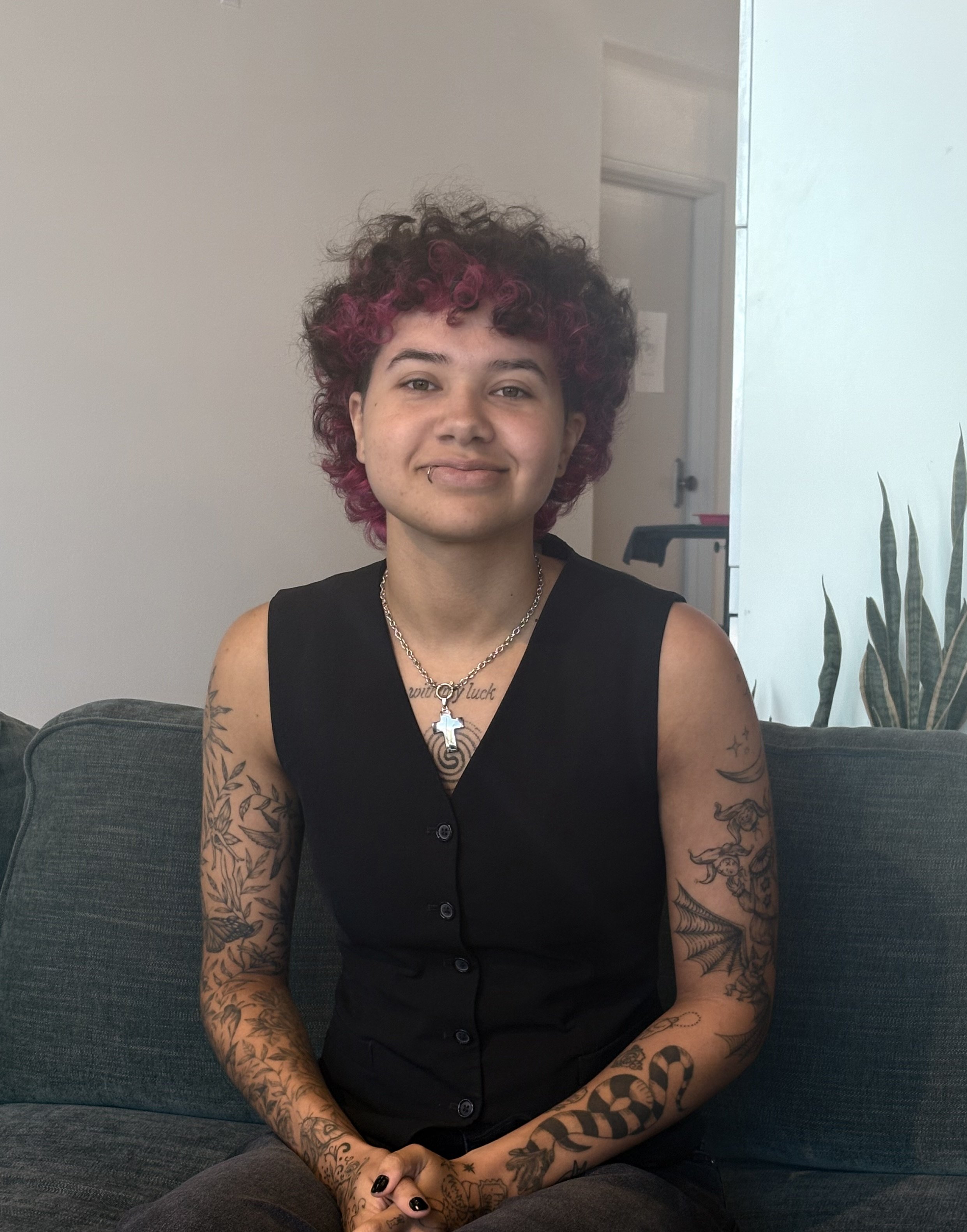 A young person with short, curly black hair with purple highlights, wearing a black sleeveless top, jewelry, and tattoos on their arms, sitting on a dark gray couch in a room with a neutral wall, a door, and plants.