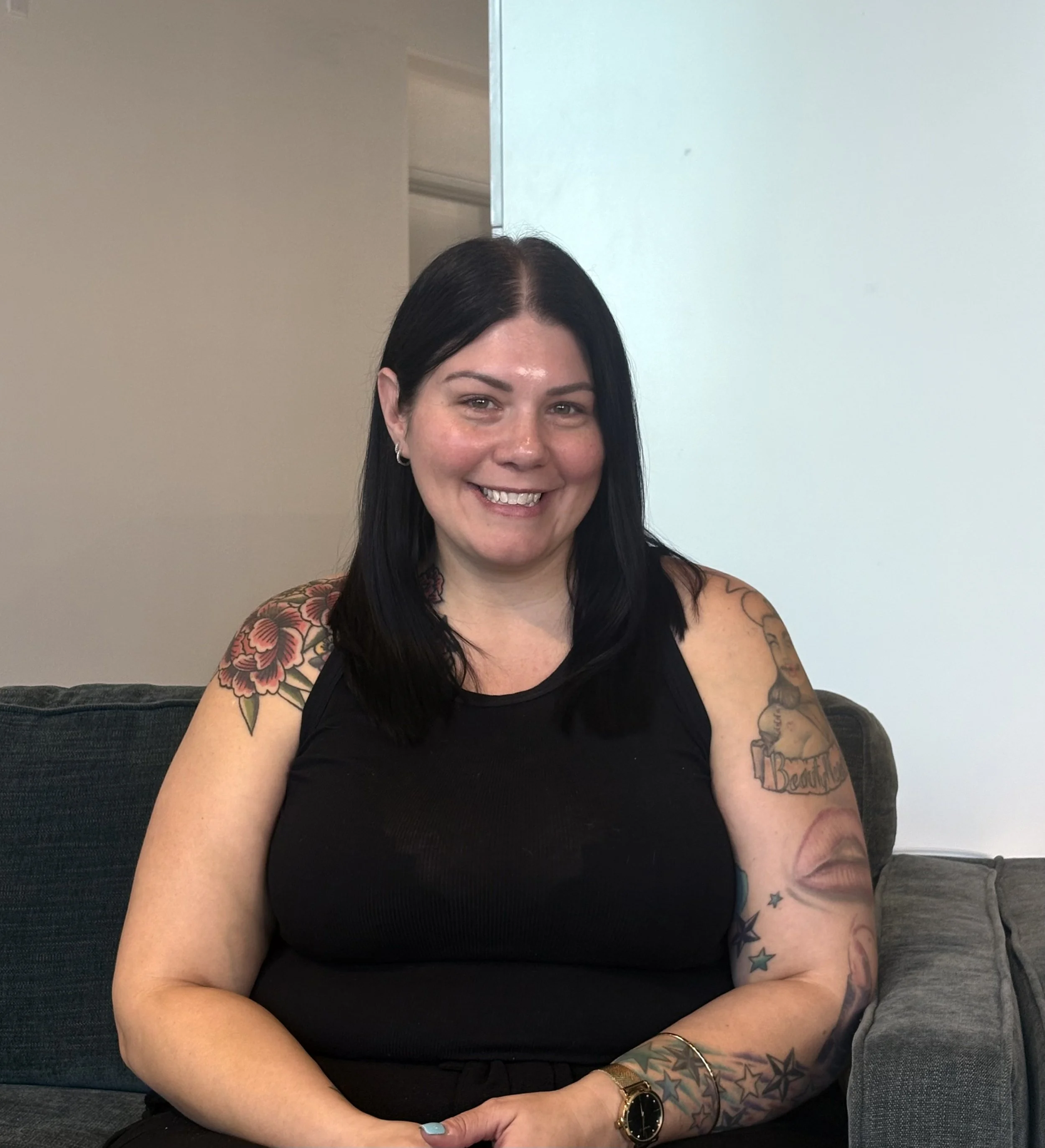 A woman with dark hair and tattoos smiling, sitting on a gray couch, wearing a black top, with colorful star tattoos on her right arm and floral tattoos on her left shoulder.