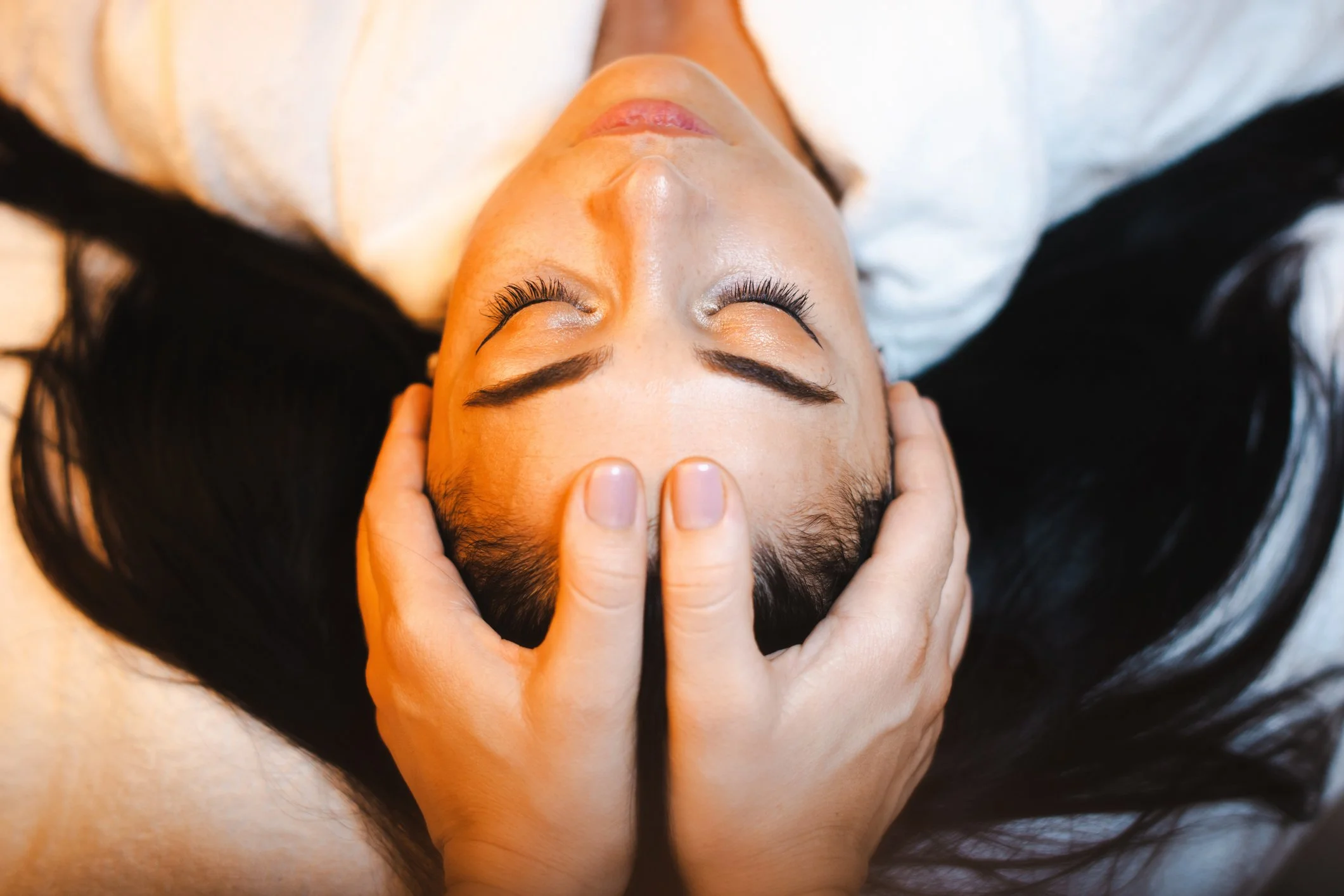 Close-up of a woman with long black hair receiving a facial massage, her eyes closed, hands gently massaging her forehead.