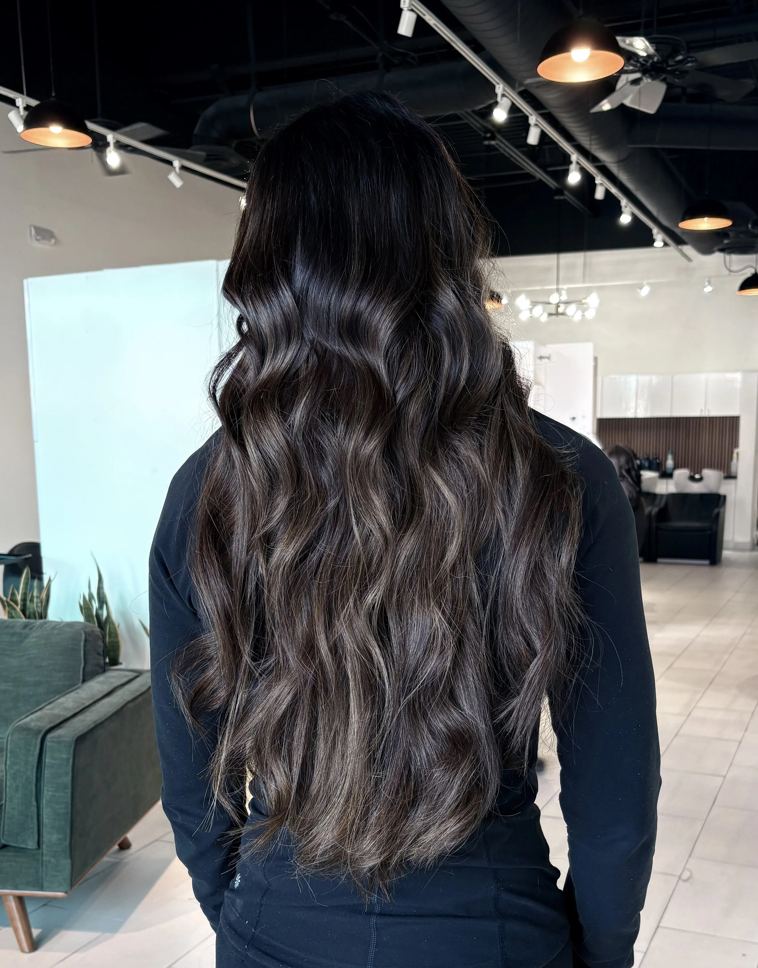 Back view of a woman with long wavy brunette hair in a salon or modern indoor space.