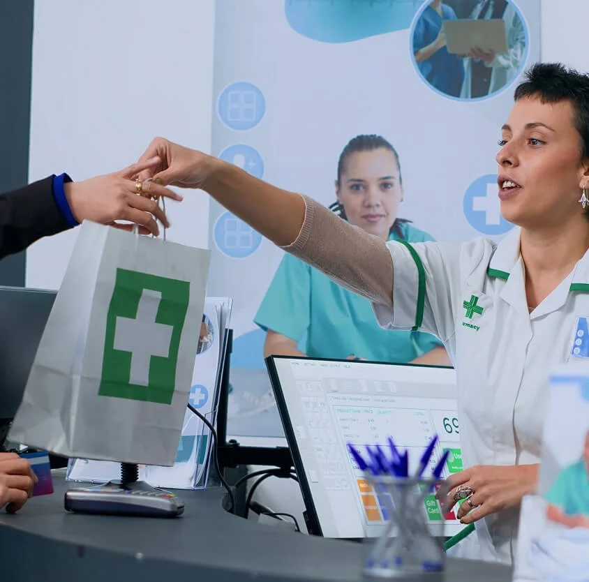 A healthcare worker in a white uniform hands a gift bag with a green cross symbol to a customer in a medical setting. In the background, another healthcare worker in teal scrubs looks on, with a computer and medical supplies on the desk.
