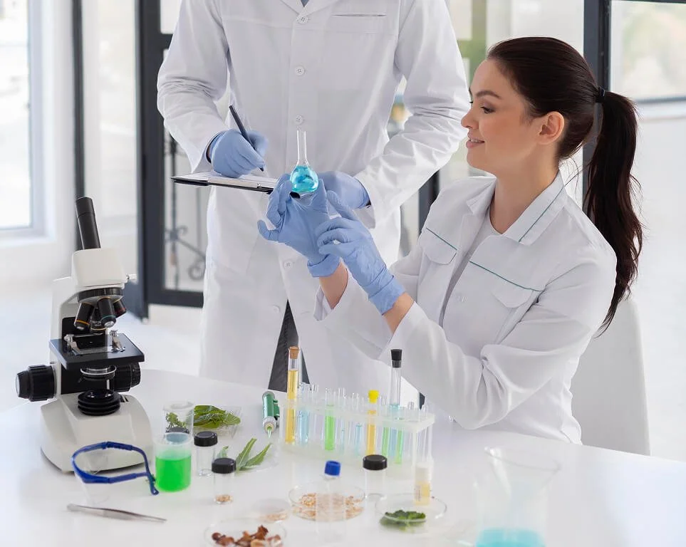 A female scientist in a lab coat and gloves holds a conical flask with a blue liquid, while a male scientist stands beside her with a clipboard, in a laboratory with microscopes and test tubes.