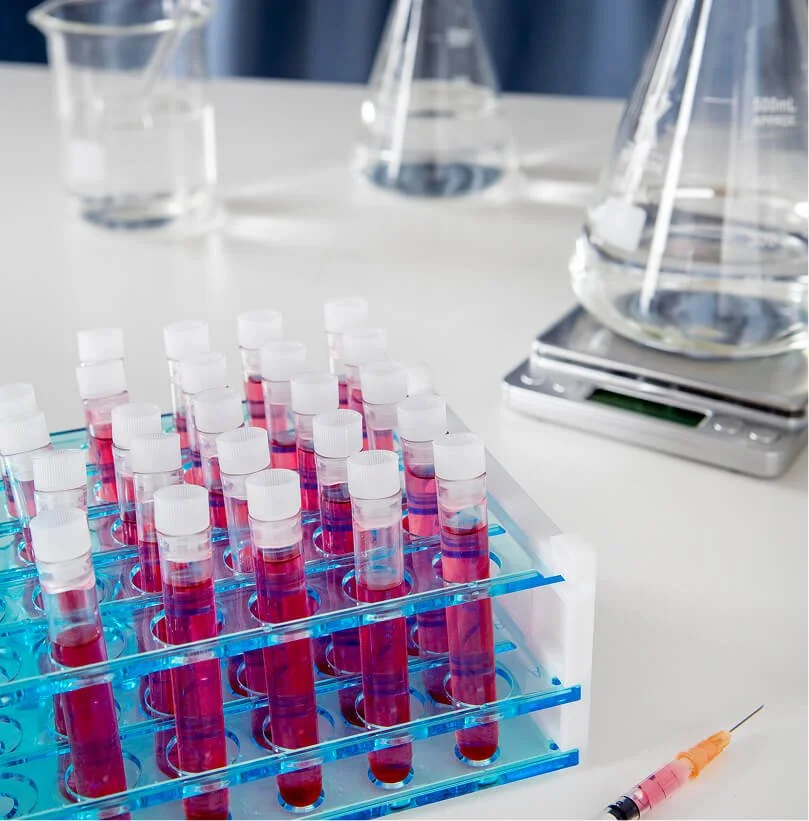 Test tubes filled with red and blue liquids in a rack, a syringe nearby, and laboratory glassware on a white table.