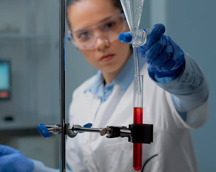 A scientist in a lab coat and safety glasses conducts an experiment using a glass pipette to transfer red liquid into a test tube, with laboratory equipment in the background.