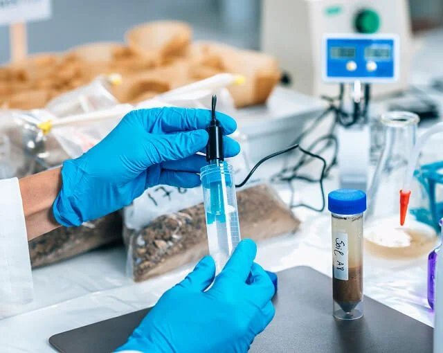 A scientist wearing blue gloves handling a syringe with a blue liquid in a laboratory setting, surrounded by lab equipment and samples.