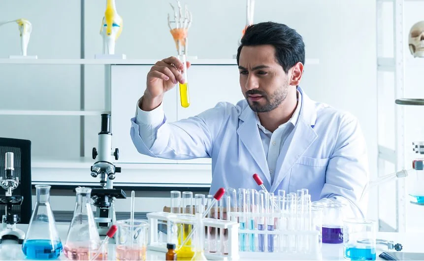 A scientist wearing a white lab coat examining a test tube with yellow liquid in a laboratory filled with various glassware and scientific equipment.