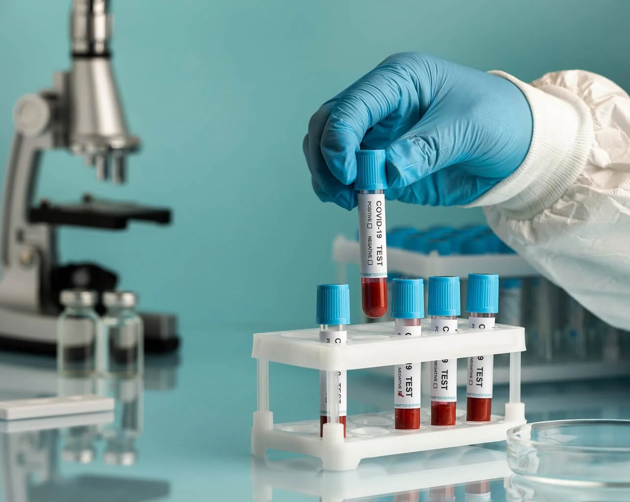 Medical professional handling a test tube labeled 'COVID-19 TEST' containing a blood sample, with additional test tubes in a rack and laboratory equipment in the background.