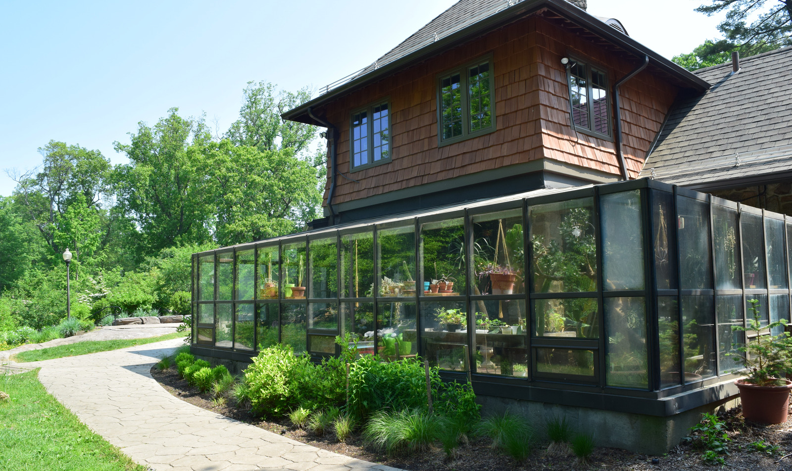 A house with a greenhouse attached, surrounded by green trees and plants on a bright, sunny day.