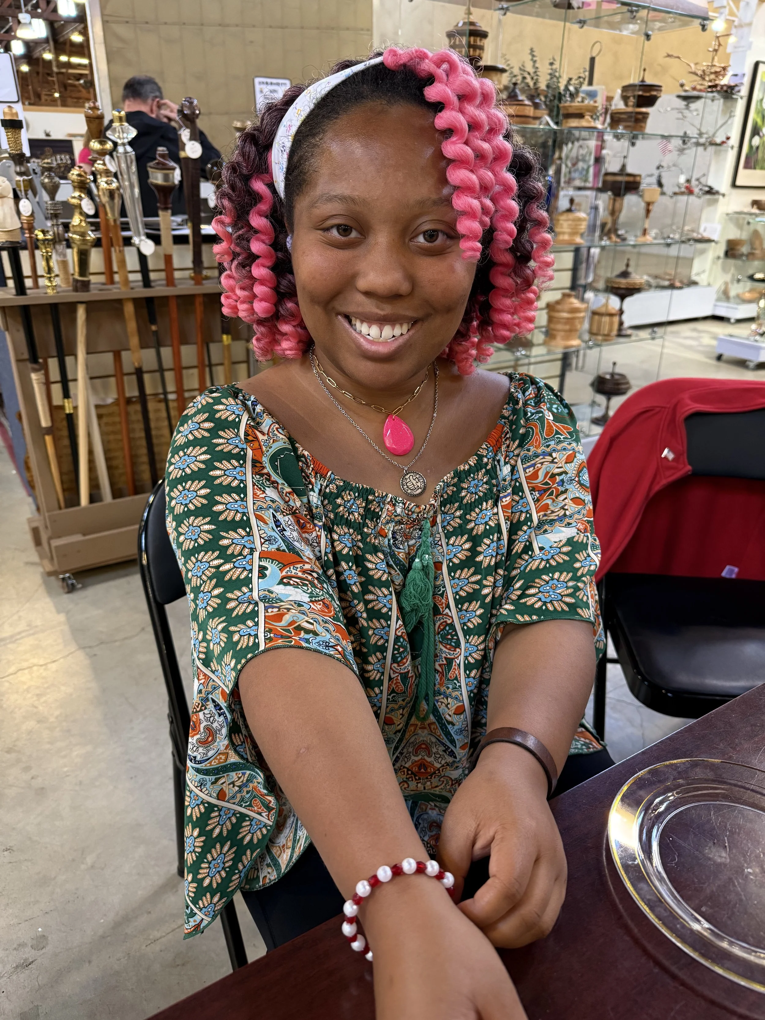 A young woman with pink curly hair, wearing a colorful patterned blouse, smiling, and holding her arm with a pearl bracelet in a store that sells wooden and glass decorative items.