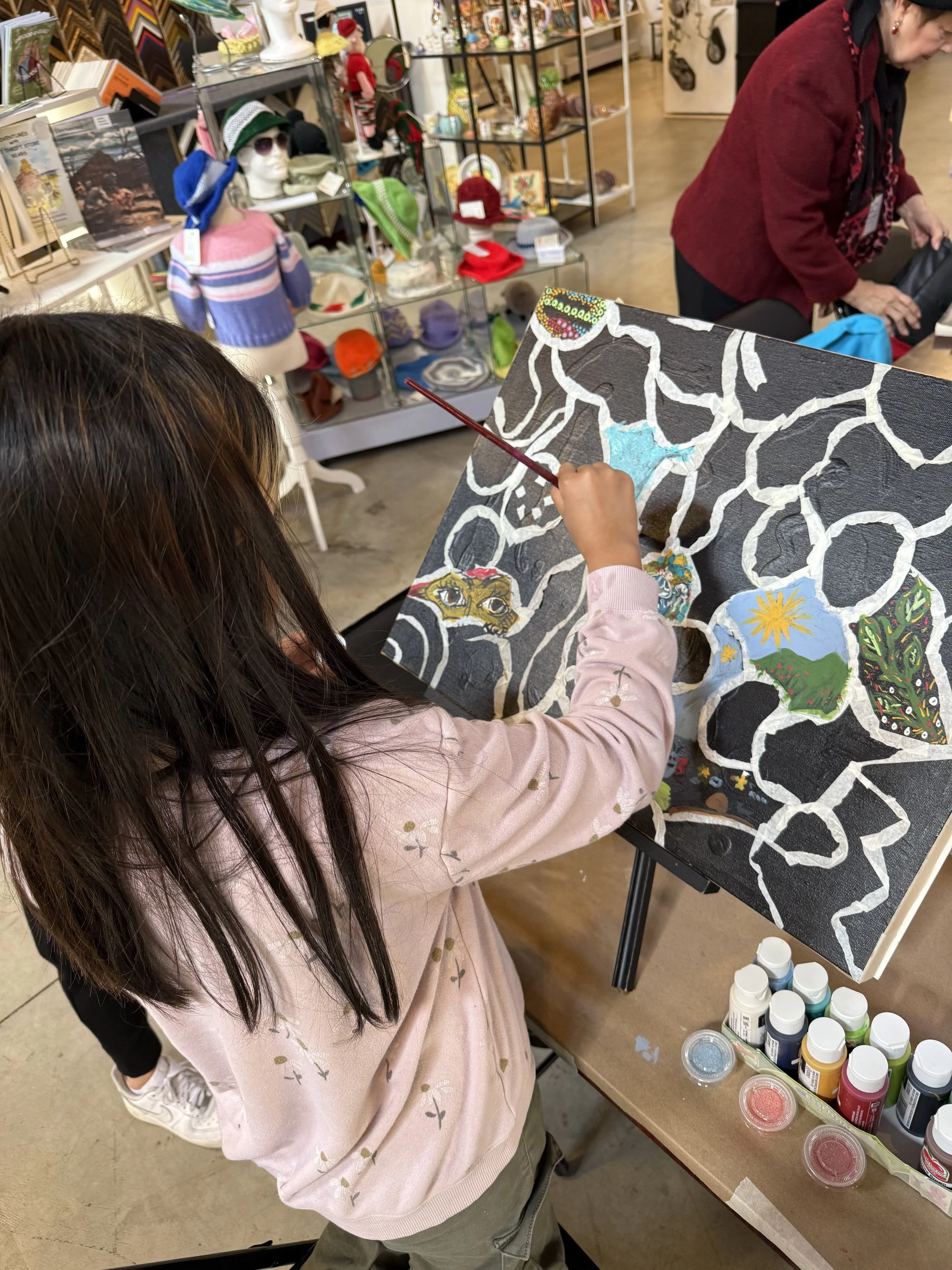 A young girl painting a colorful abstract design on a black canvas in an art store or workshop, with bottles of paint and other supplies on the table.