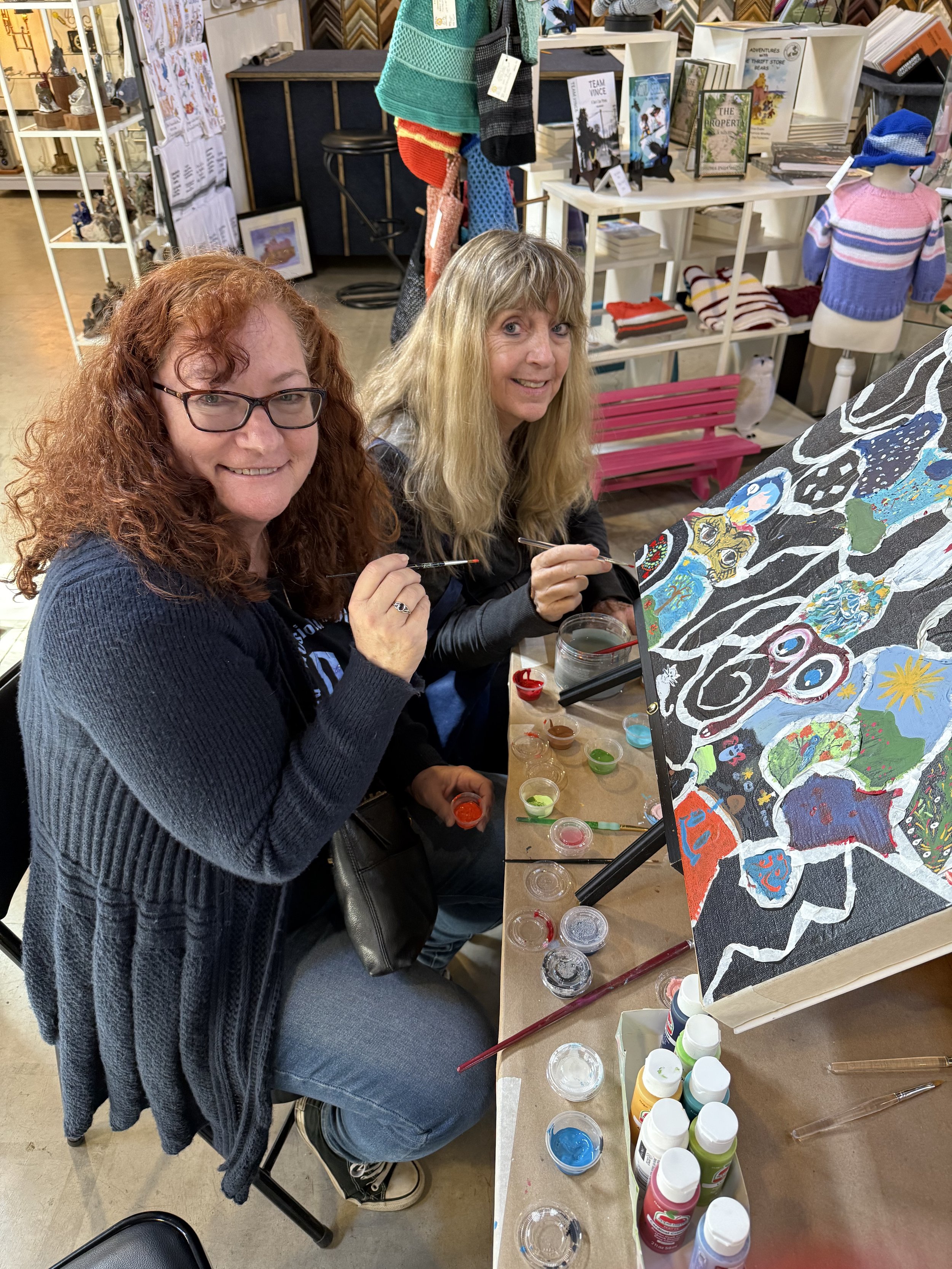 Two women sitting at a table painting a colorful, abstract artwork on a canvas in a craft store.