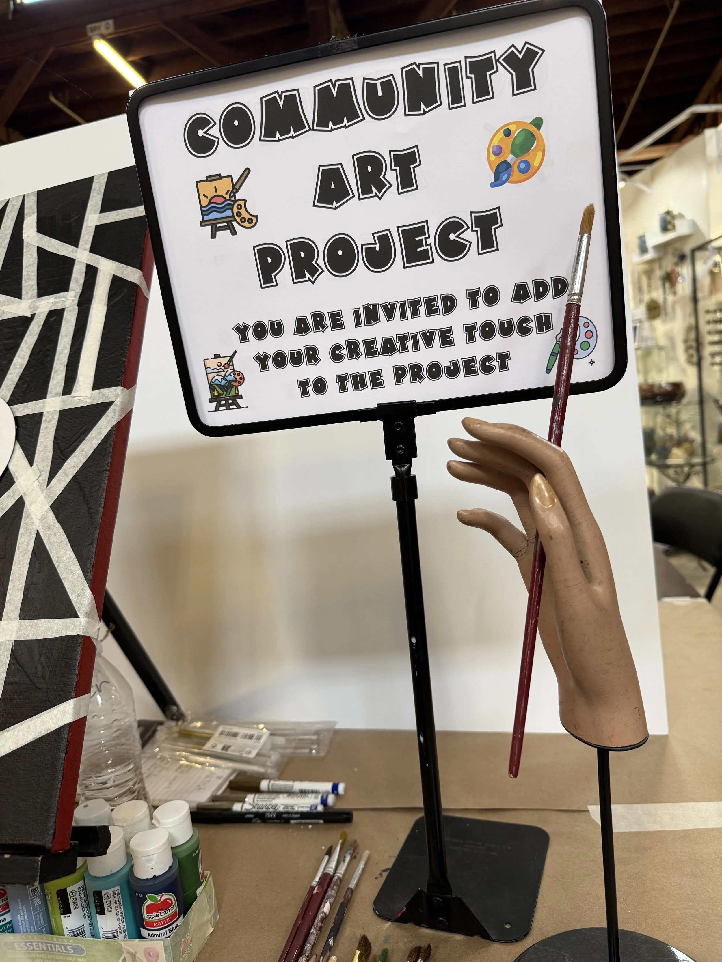 Sign for a community art project inviting people to add their creative touch, decorated with small illustrations of an artist's palette and brushes.