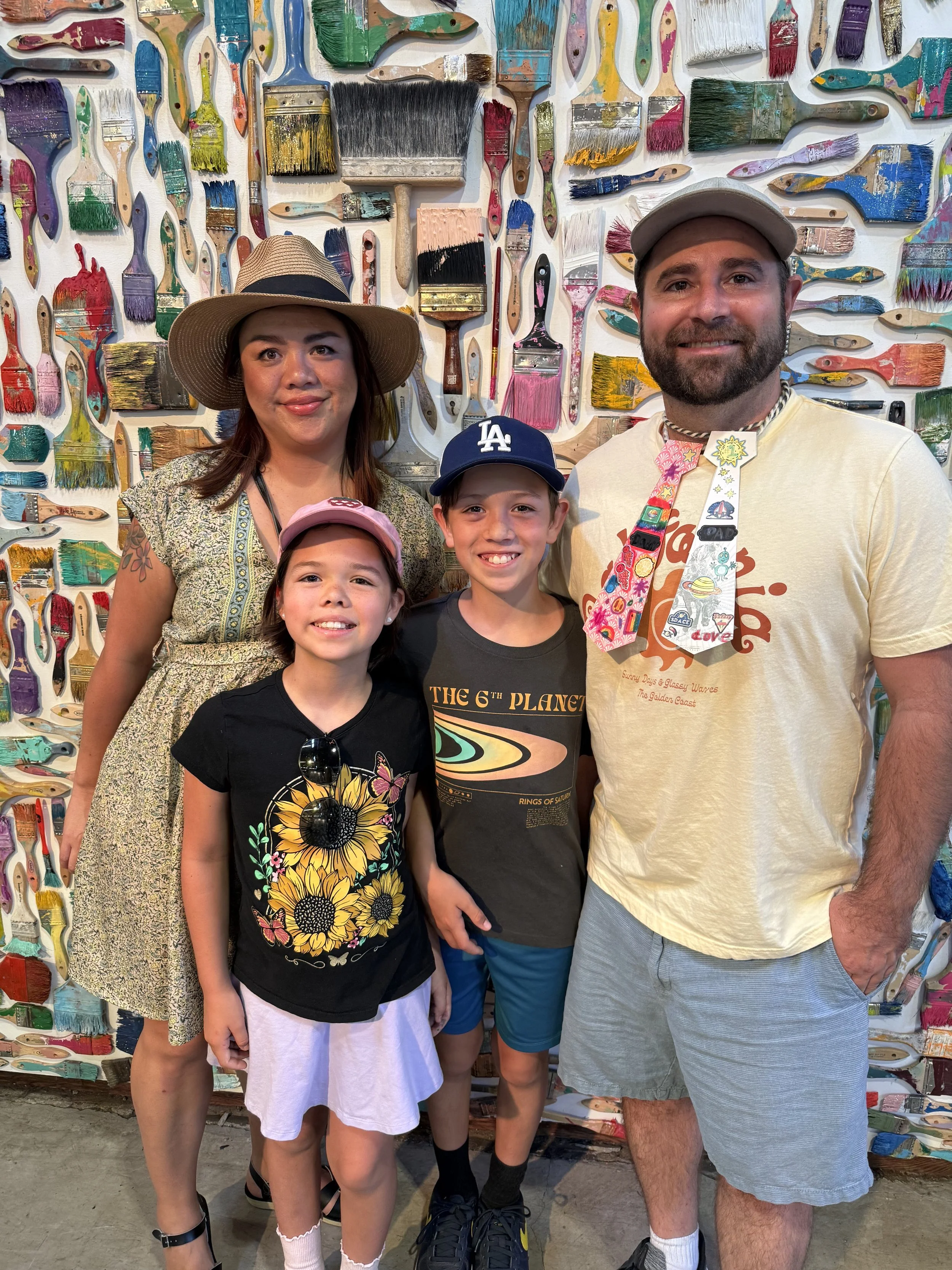 A family of five posing in front of a colorful wall decorated with numerous paintbrushes. The woman on the left wears a wide-brimmed hat and a patterned dress. The man on the right has a beard, wears a gray cap, a yellow T-shirt, and a colorful necktie. There are two children in between: a girl with a pink cap, black T-shirt with sunflowers and butterflies, and white skirt; a boy with a navy baseball cap, gray T-shirt with space graphic, and blue shorts.