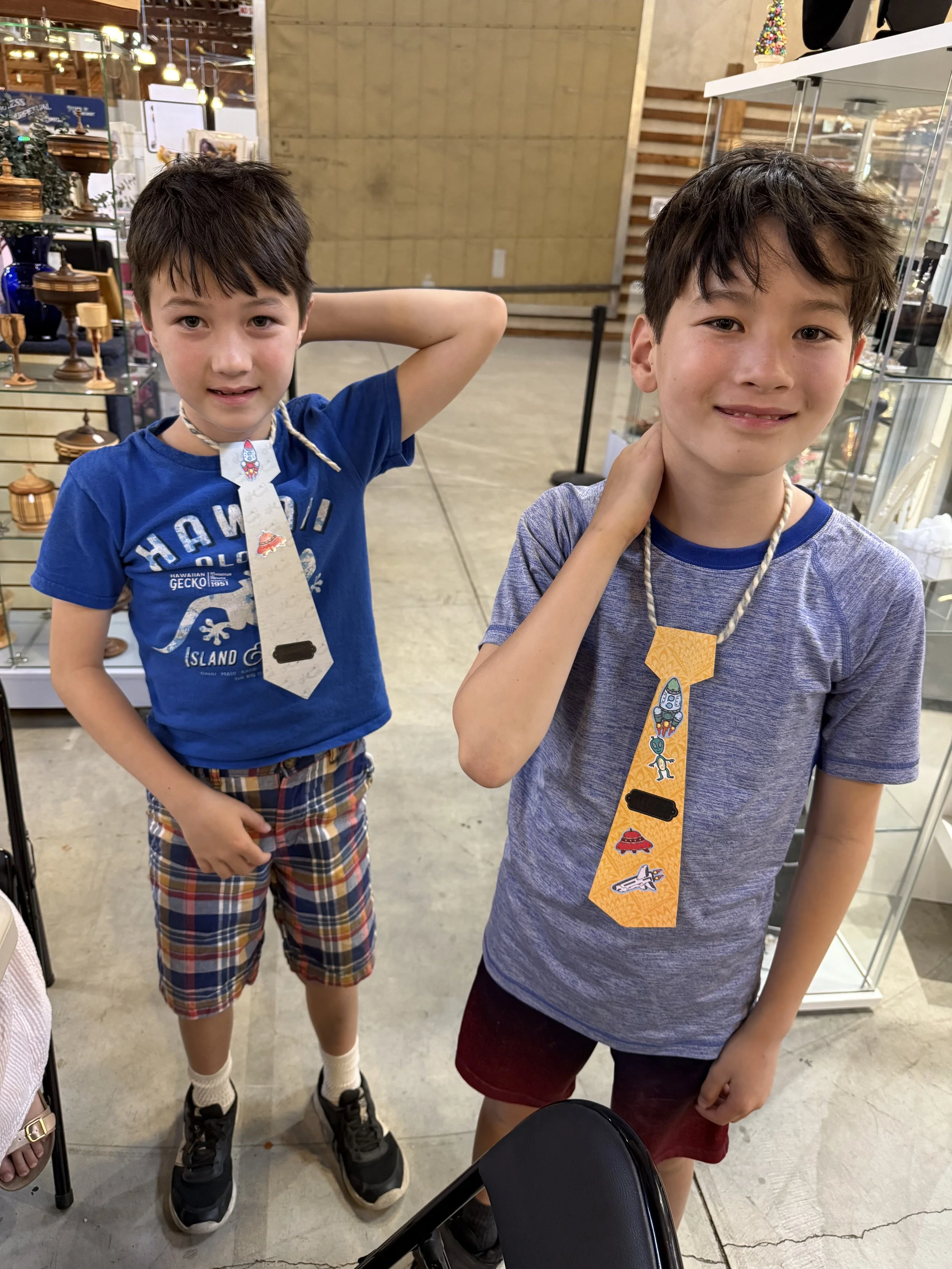 Two young boys standing inside a store, wearing handmade paper ties around their necks decorated with colorful stickers and drawings, smiling at the camera.