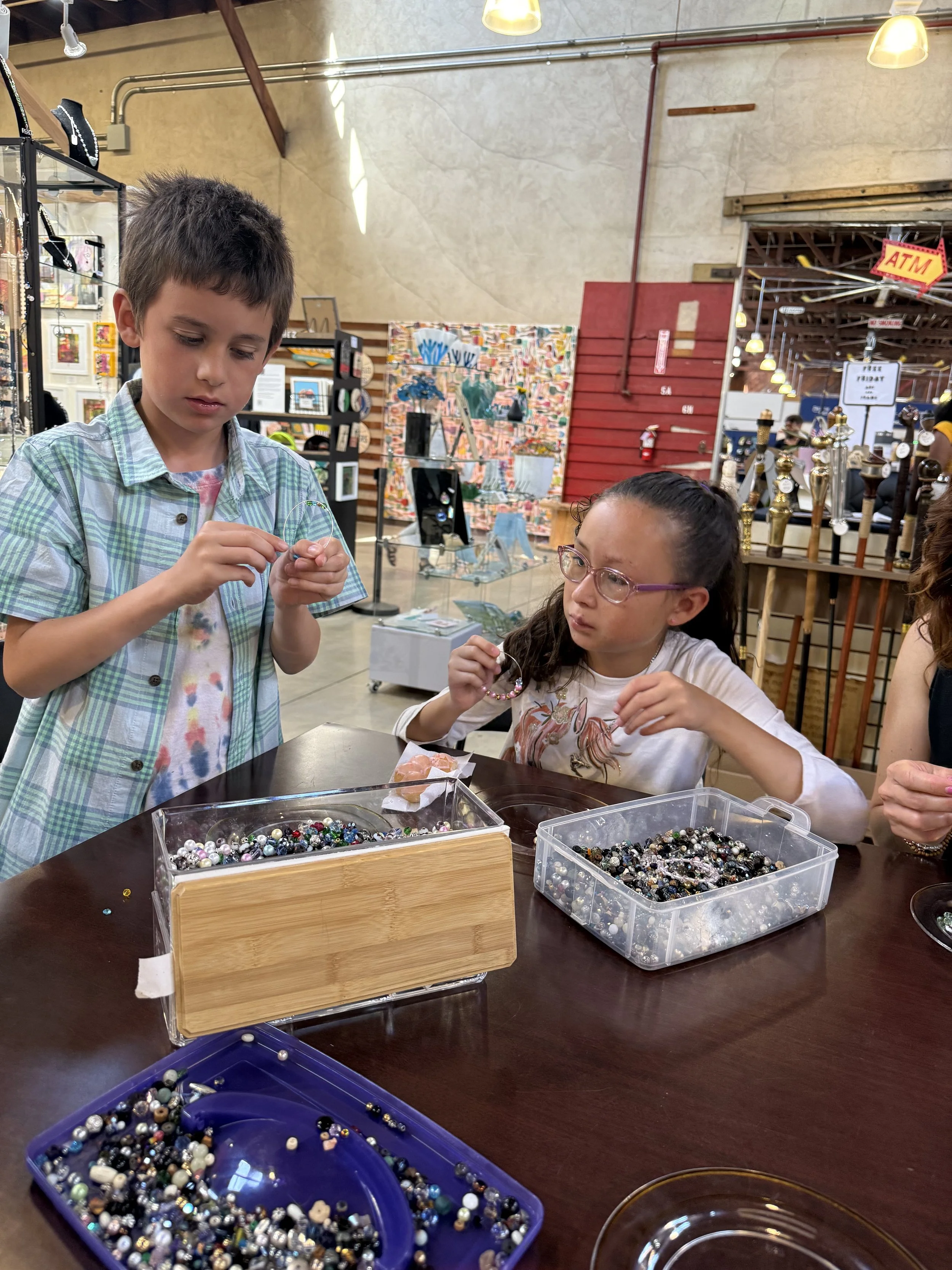 Two children, a boy and a girl, sit at a table in a store, creating jewelry with colorful beads. The boy is focused on threading beads, while the girl, wearing glasses, looks at her bracelet. There are multiple containers of beads on the table.