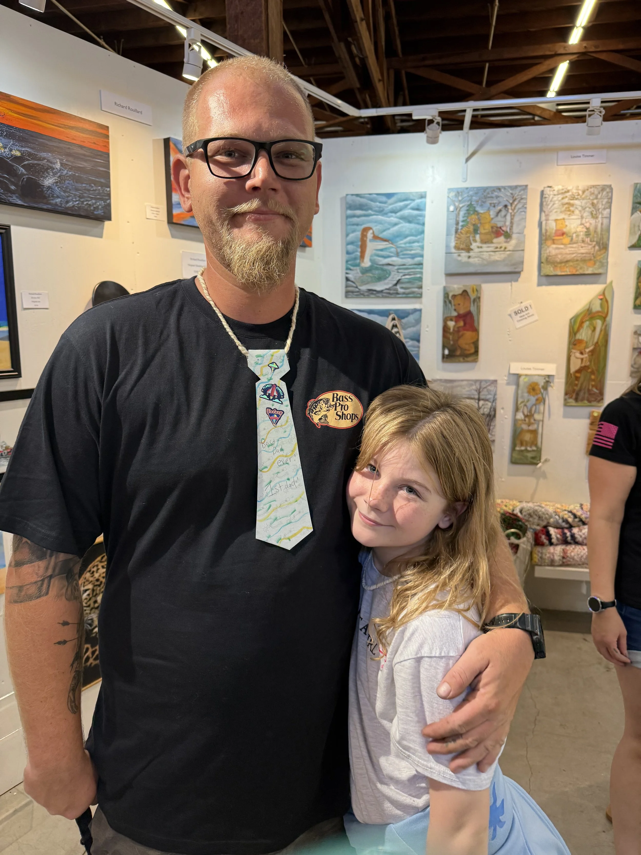 A man with glasses, a beard, and a mustache standing next to a young girl with long blonde hair, both smiling inside an art gallery. The man is wearing a black Bass Pro Shops shirt and a handmade tie with colorful writing, and the girl is wearing a light gray shirt. Artwork is displayed on the walls behind them.