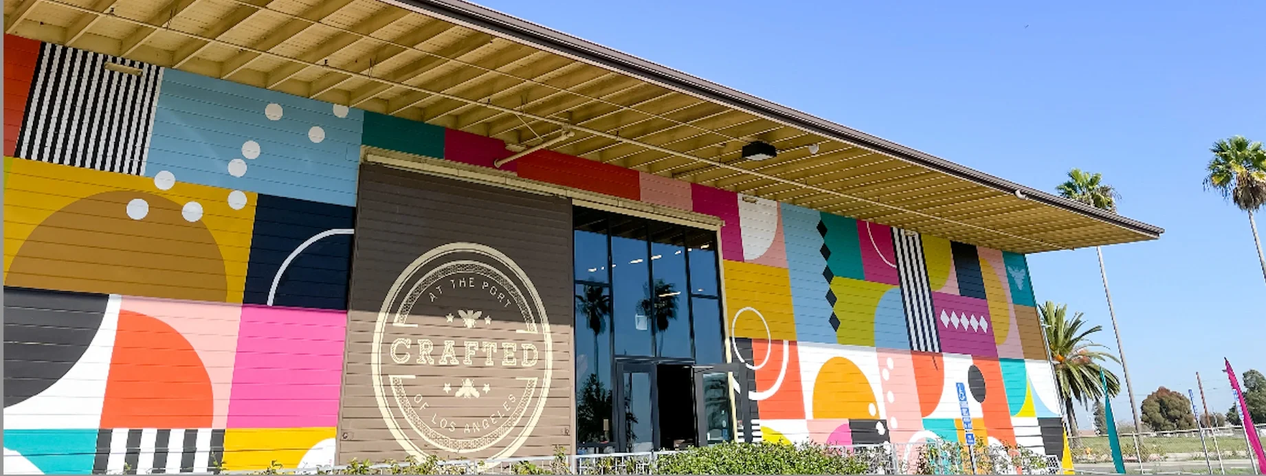 Colorful mural on a building exterior with geometric shapes and patterns, with a sign reading "At The Port Crafted Los Angeles" at the entrance, and palm trees in the background under a clear blue sky.