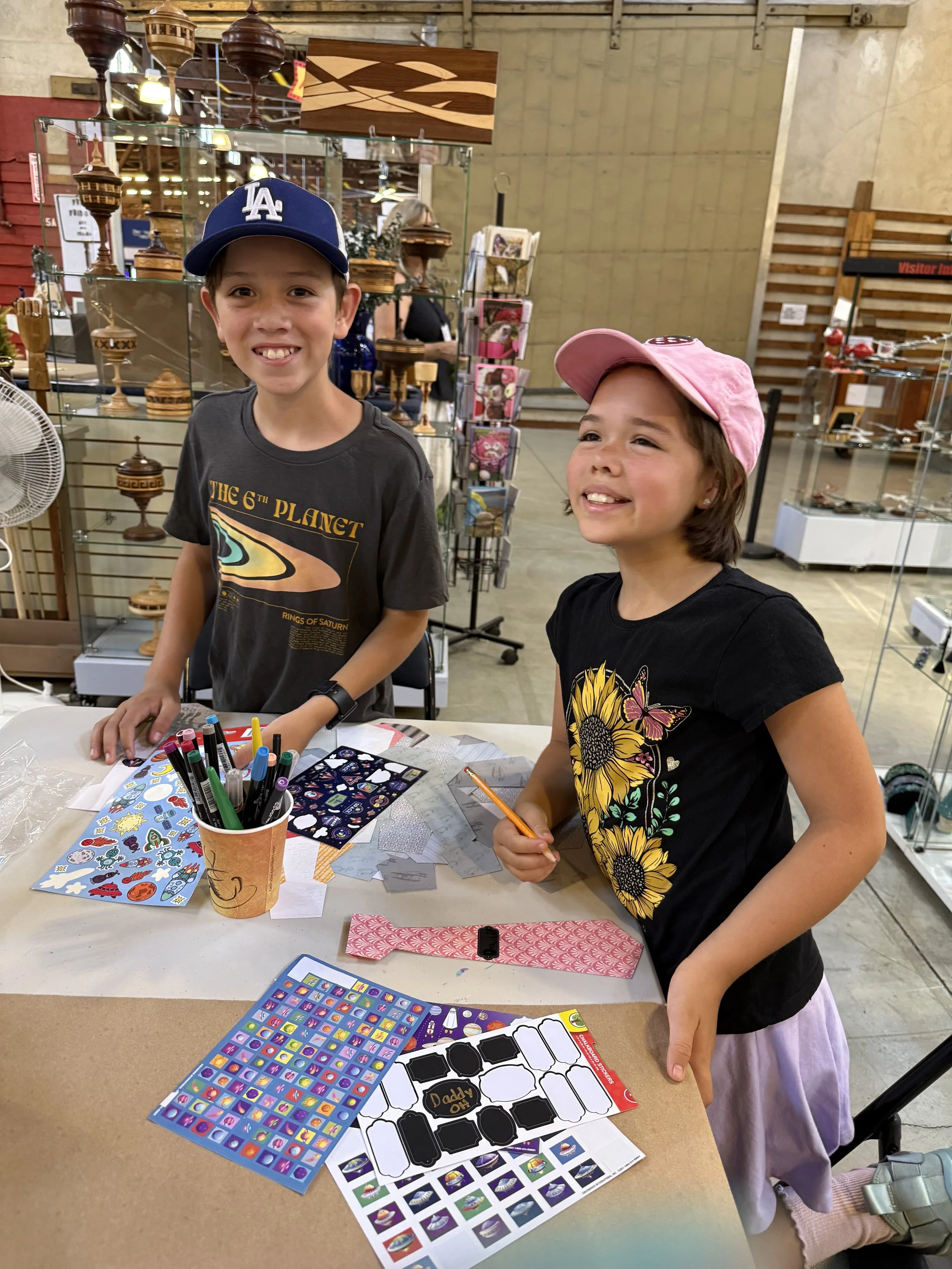 Two children, a boy wearing a blue Los Angeles Dodgers cap and a girl wearing a pink cap, smiling at a table with stickers and craft supplies, inside a store with wooden shelves and display cases.