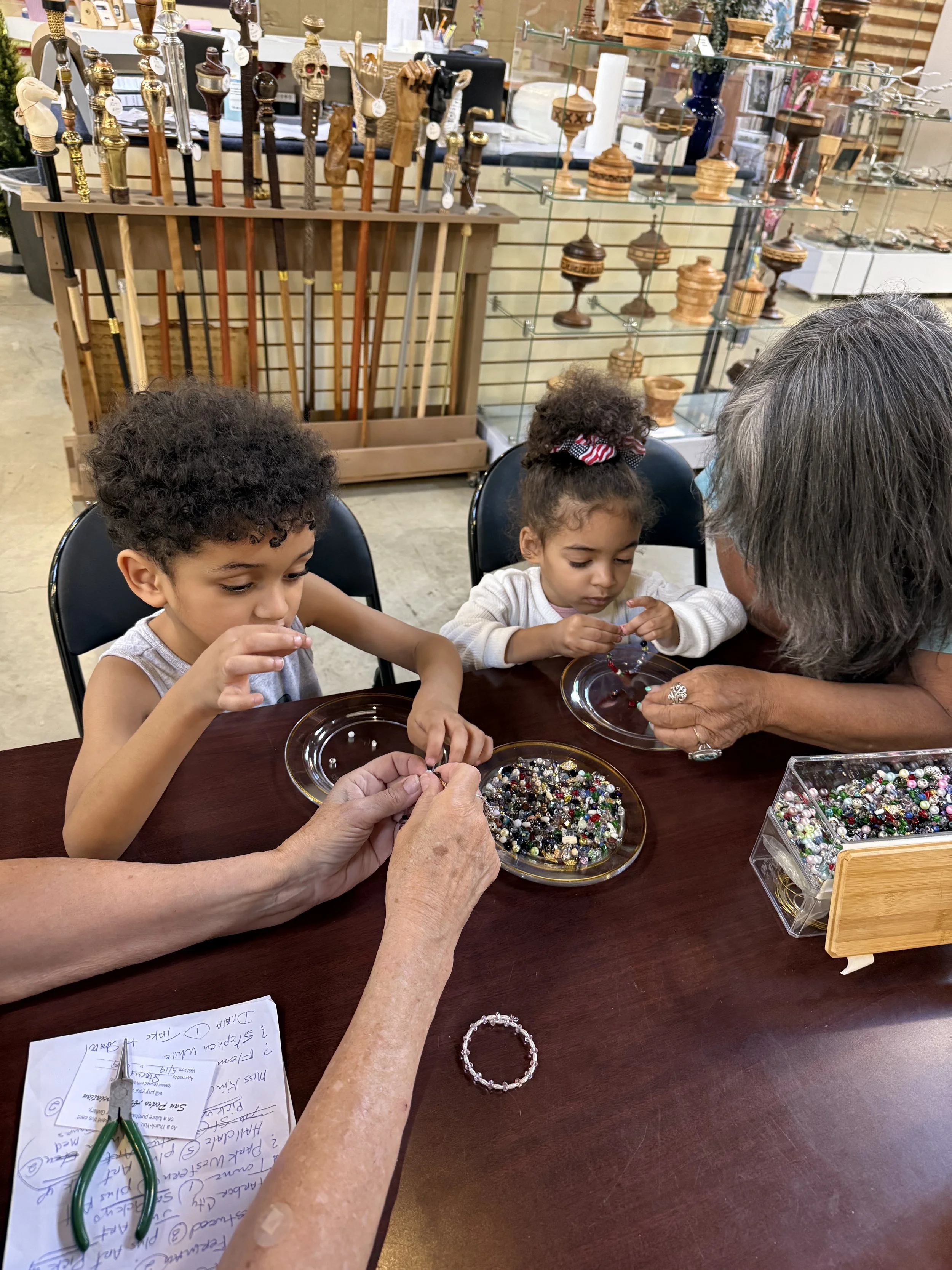 Two young girls and an elderly woman working on jewelry-making at a table, with beads and jewelry supplies around them in a craft store or workshop.