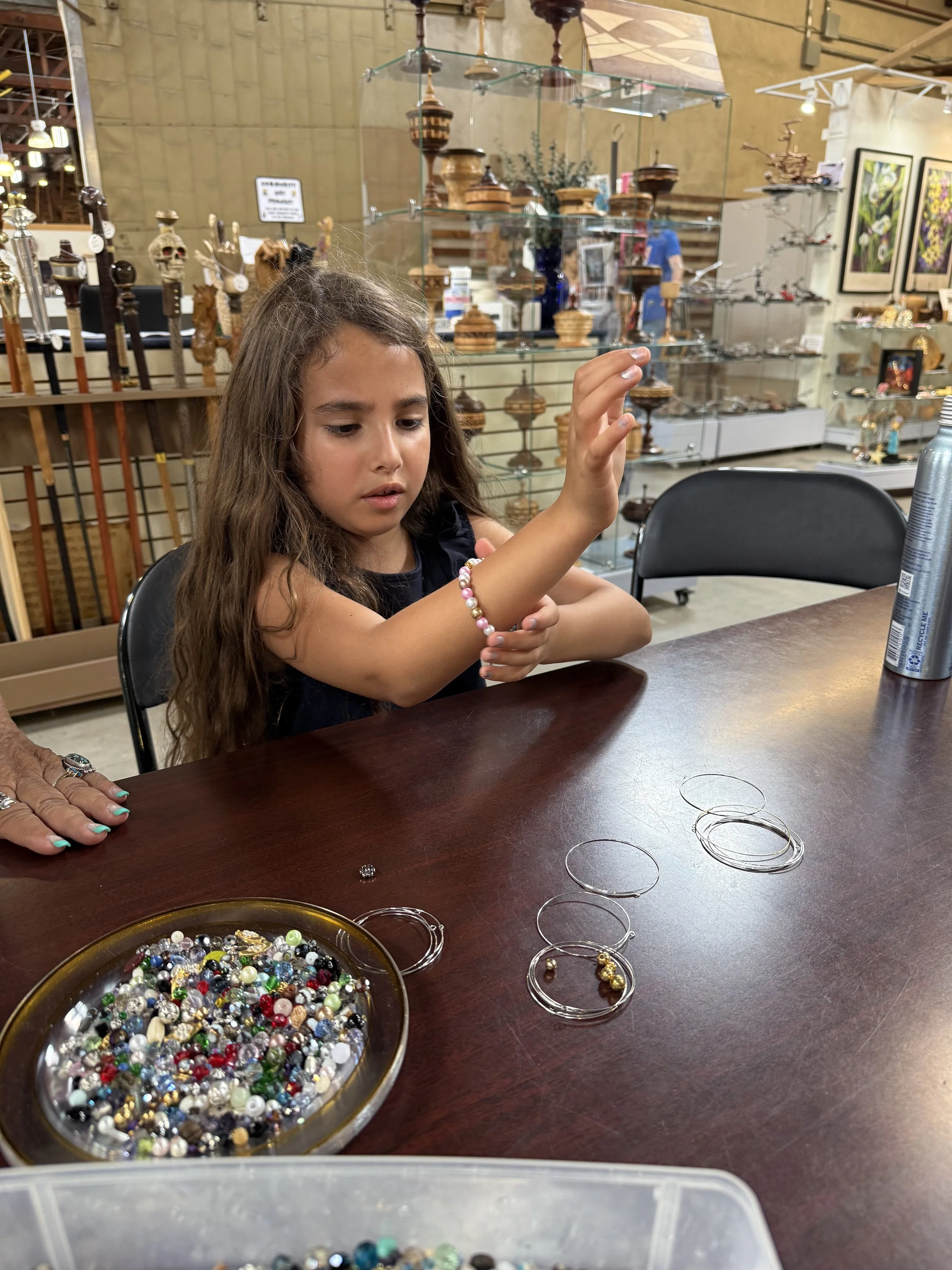 A young girl sitting at a table with a variety of bracelets and jewelry, reaching out to pick or examine a bracelet, in a store with glass display cases filled with various decorative items and art on the walls.