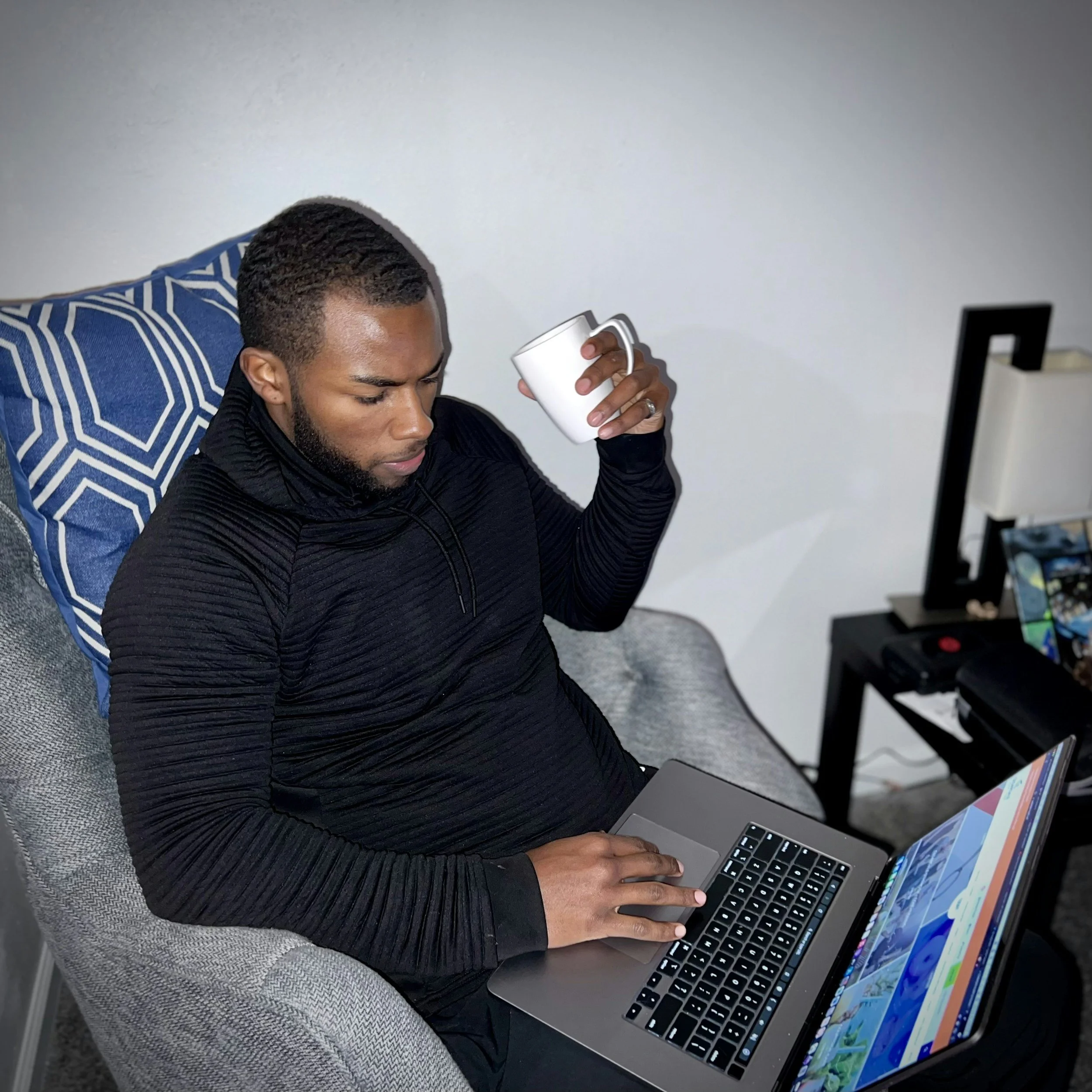 Man sitting on a gray chair, working on a laptop, holding a white mug, wearing a black long-sleeve shirt, with a blue patterned pillow behind him and a black table with a computer monitor nearby.