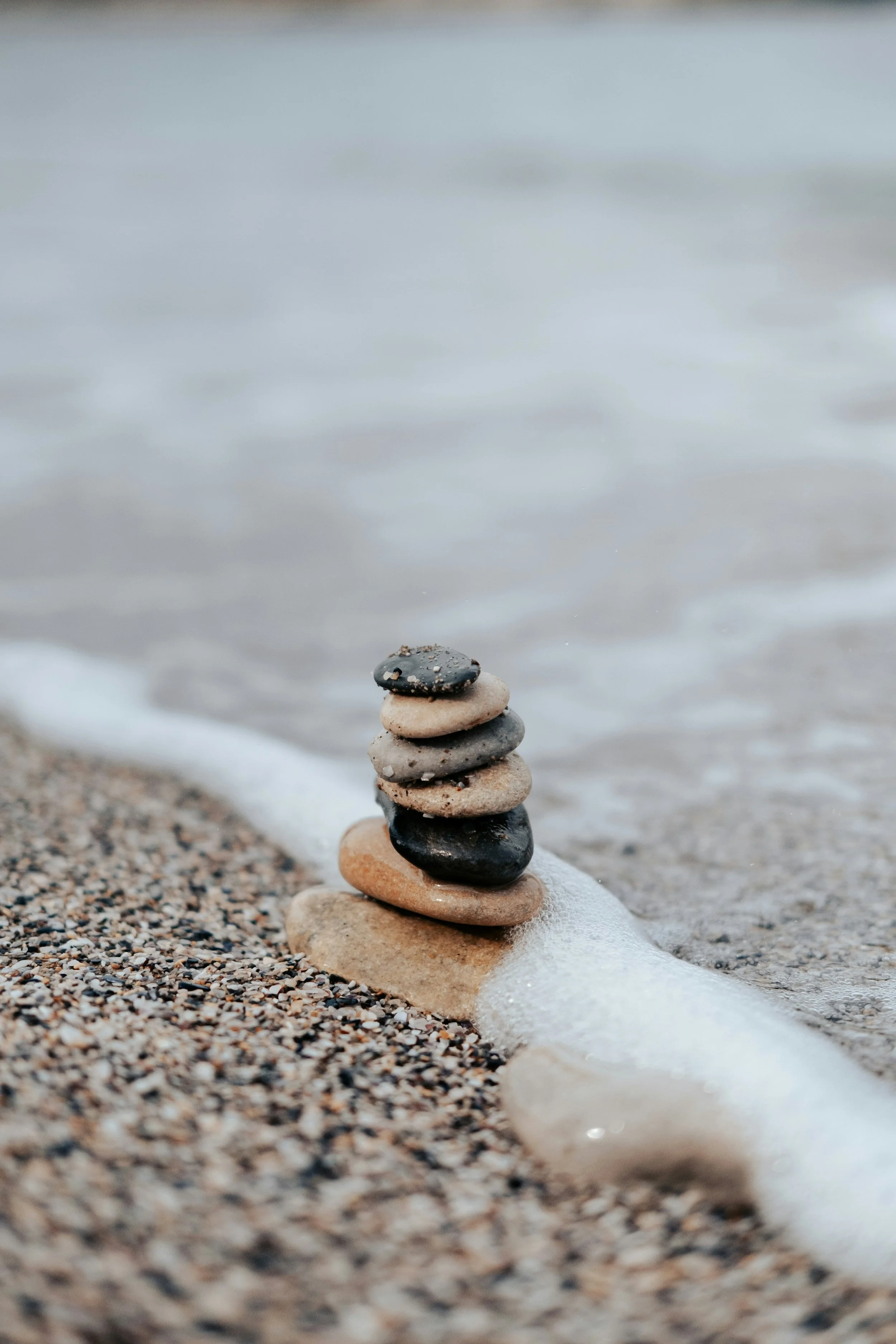 A stack of seven smooth, multicolored stones balanced on a pebble on a sandy beach, with waves and water in the background.