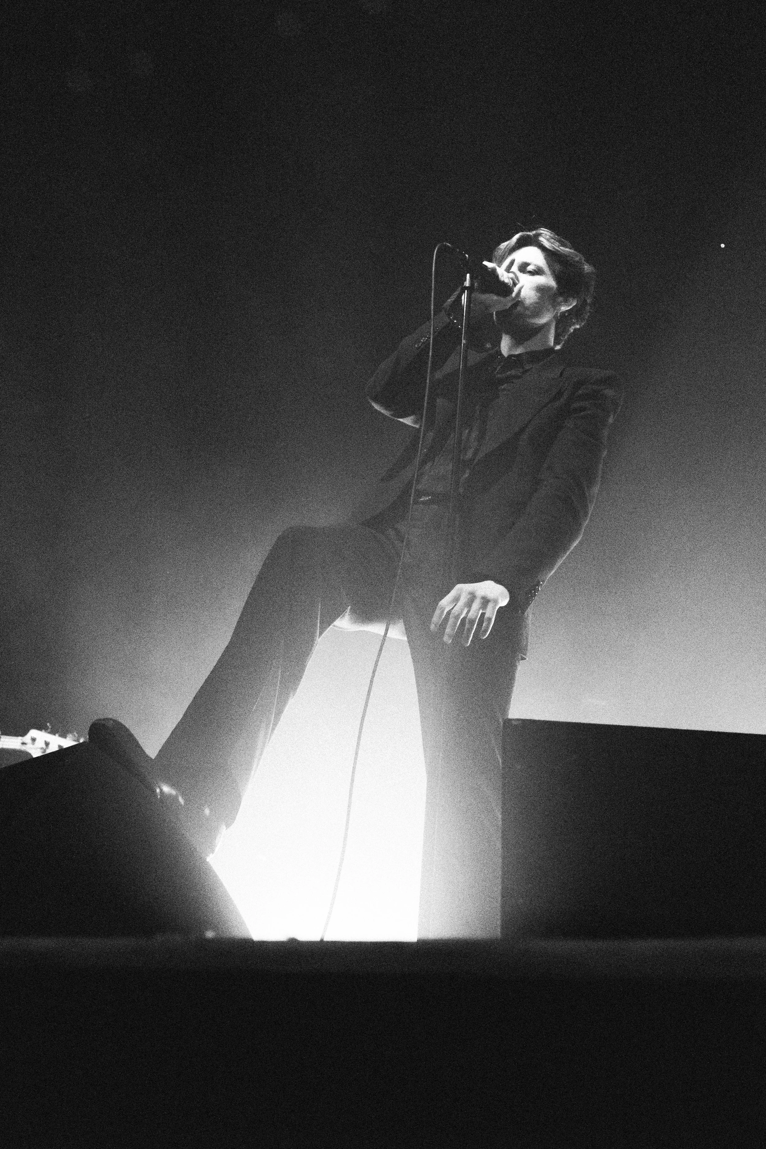 A person singing into a microphone on stage, illuminated from behind creating a silhouette effect.