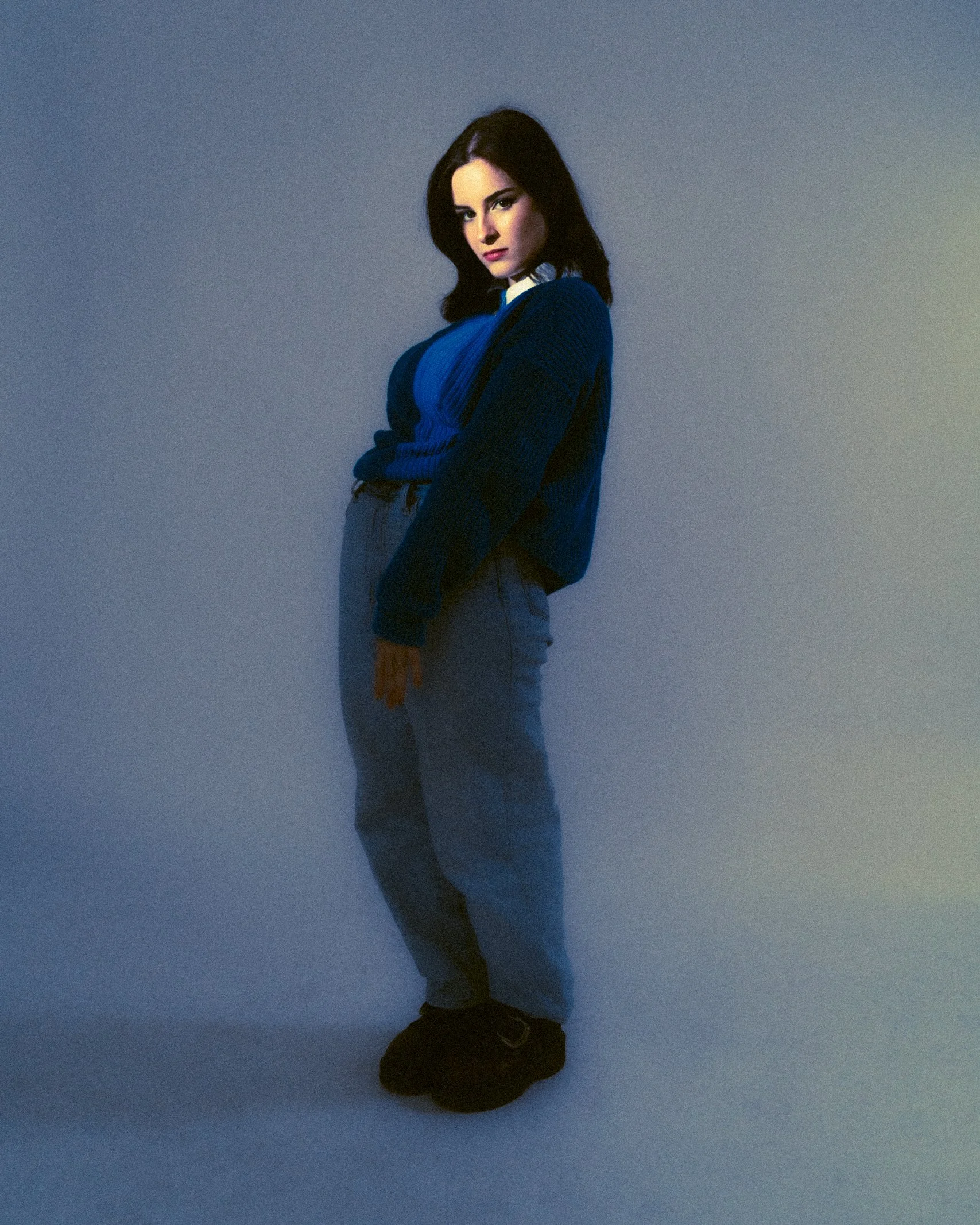 A woman with dark hair posed against a gradient background, wearing a blue sweater and gray pants.