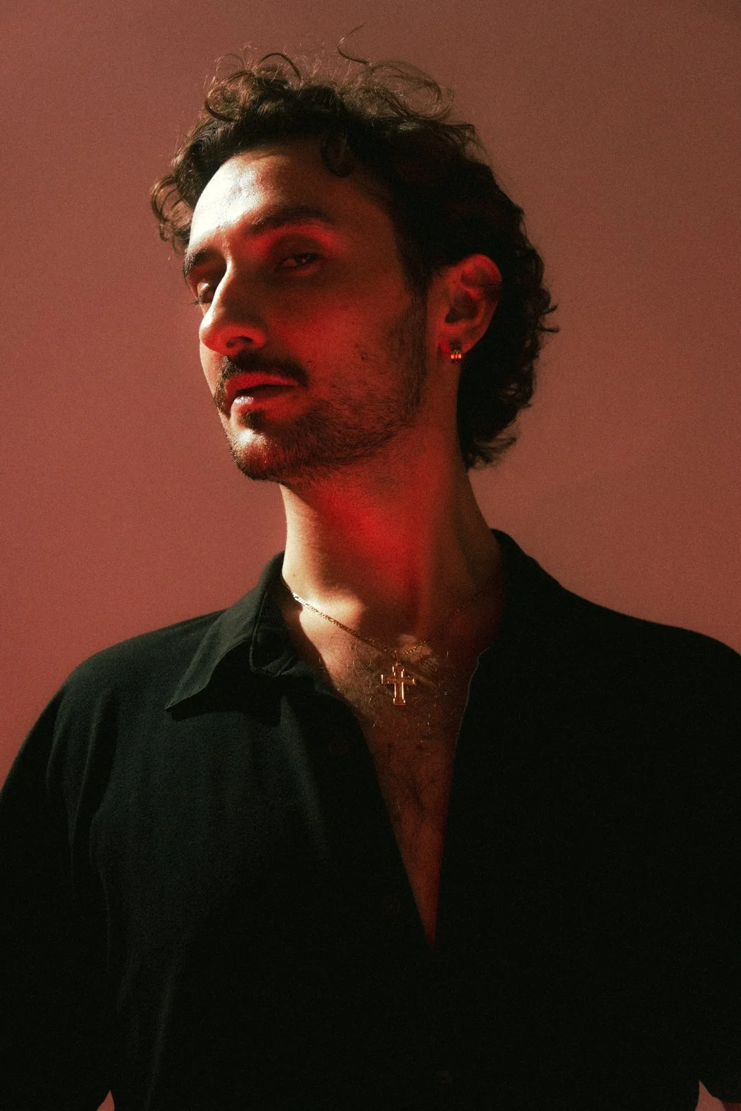 A man with dark curly hair and a beard, wearing a black shirt with the top few buttons open, a gold chain with a cross pendant, and small hoop earrings. The background is plain red, and he is looking slightly to the side with a serious expression.