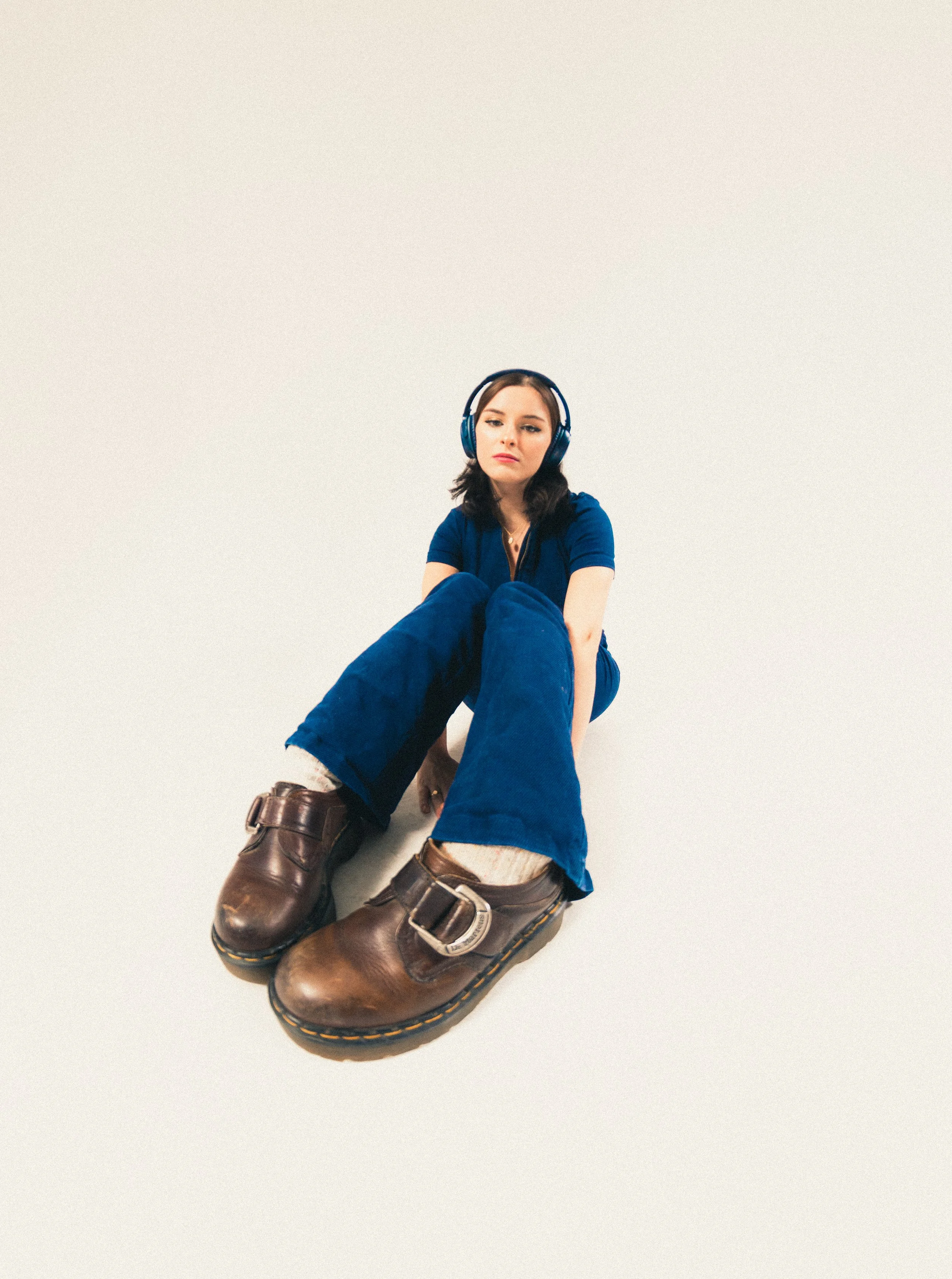 A young woman with dark hair and headphones sitting on the floor, wearing a blue outfit and large brown boots, against a plain white background.