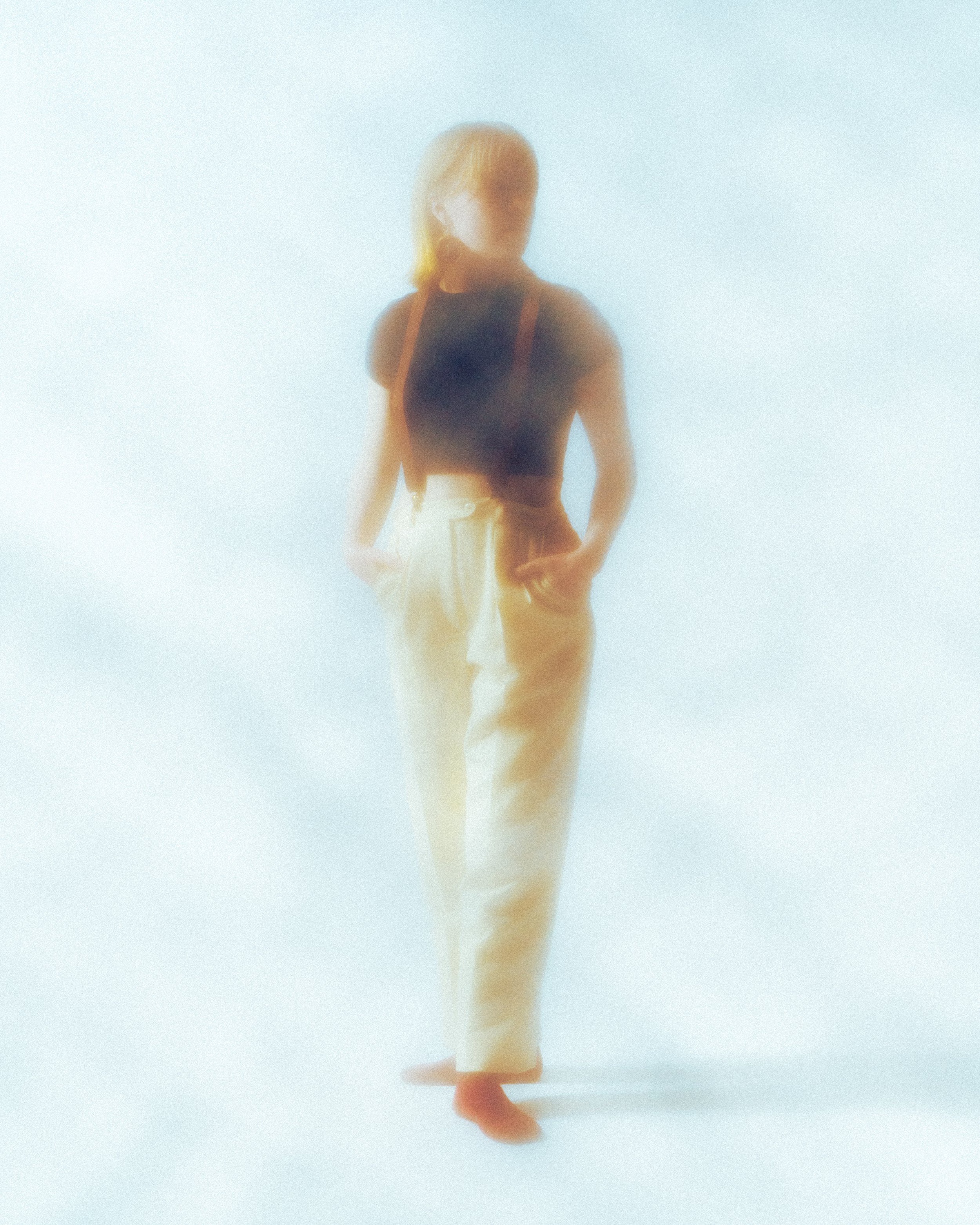 A person standing against a light background, seen from the back, wearing a black sleeveless top and beige pants, with a bag on their shoulder.