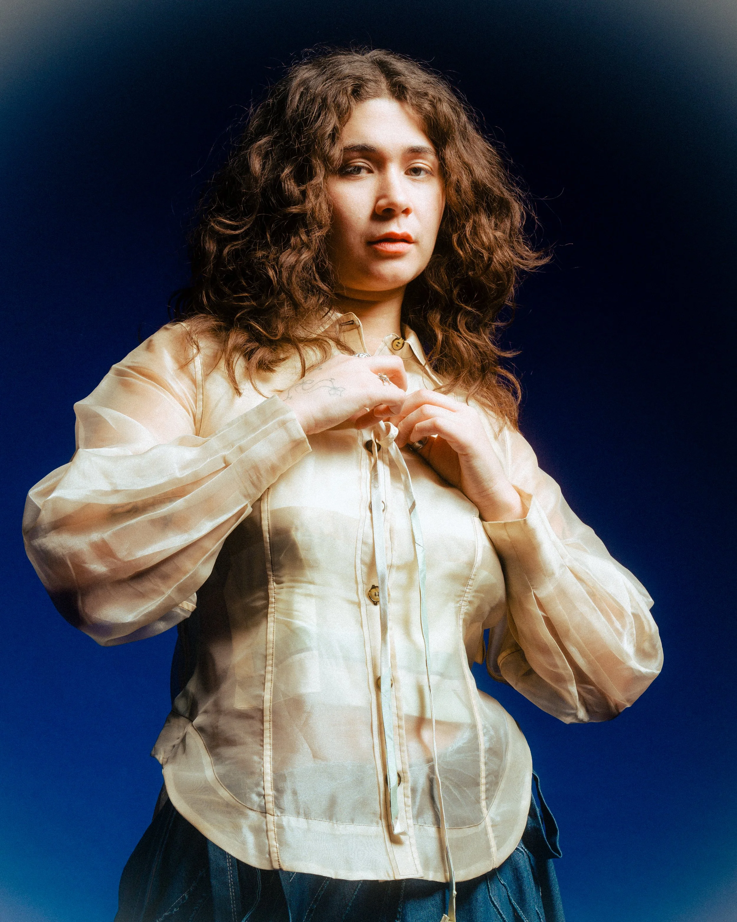 Close-up of a woman with curly hair, wearing a beige satin blouse, against a dark blue background.