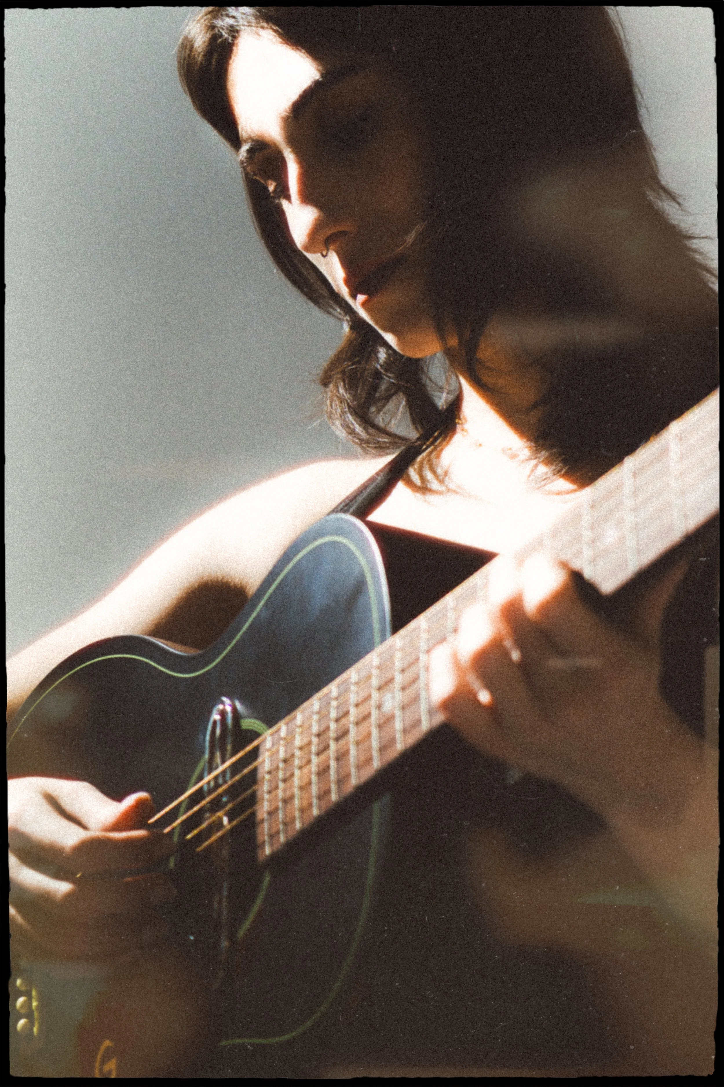 A person playing a black acoustic guitar with a wooden fretboard, focusing on their left hand on the strings and their face partially visible. They have shoulder-length dark hair and are wearing a sleeveless top. Soft lighting highlights their face a
