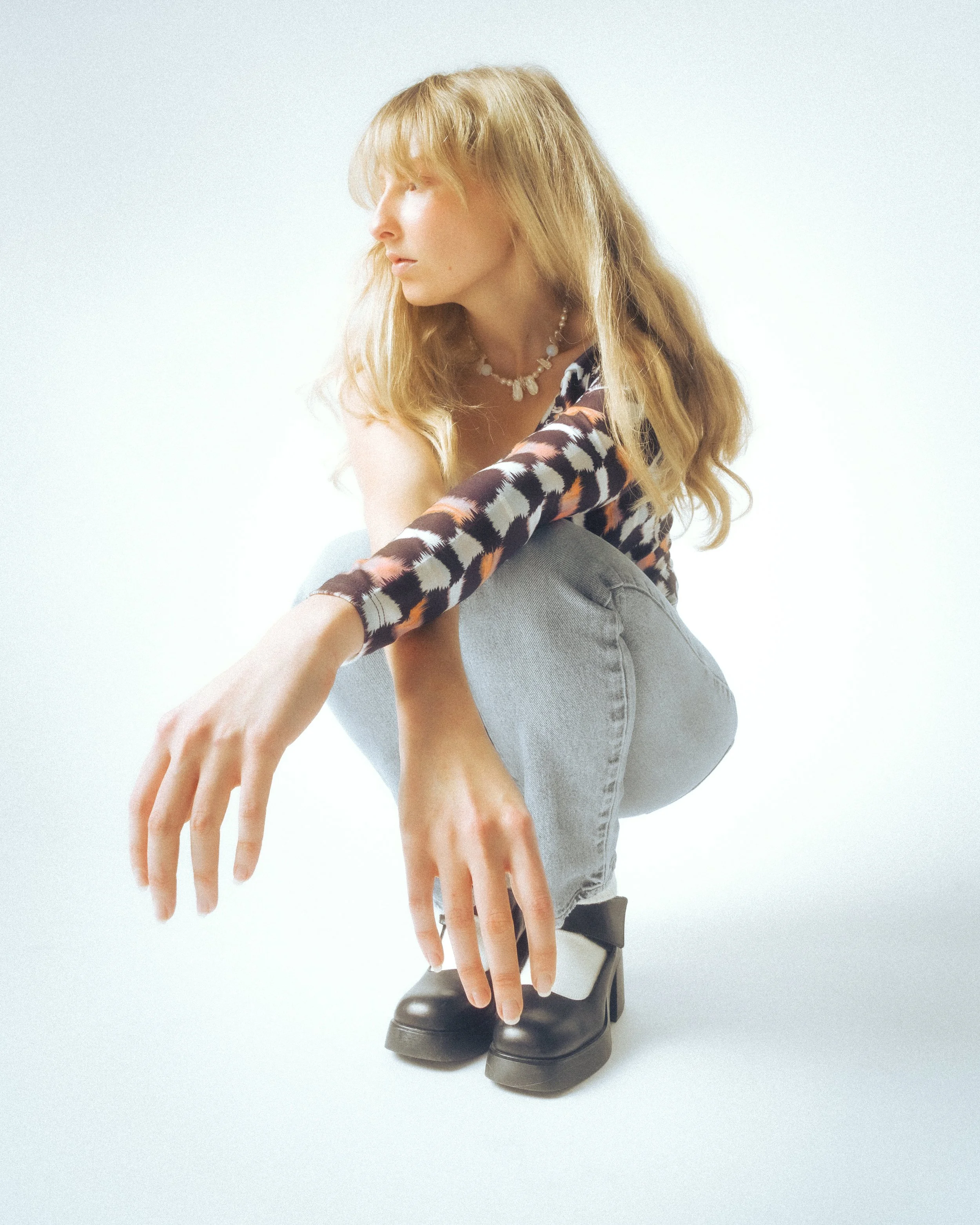 Fashion doll with blonde hair, wearing a black and orange patterned top, gray pants, black boots, and a pearl necklace, sitting with one hand extended downward.