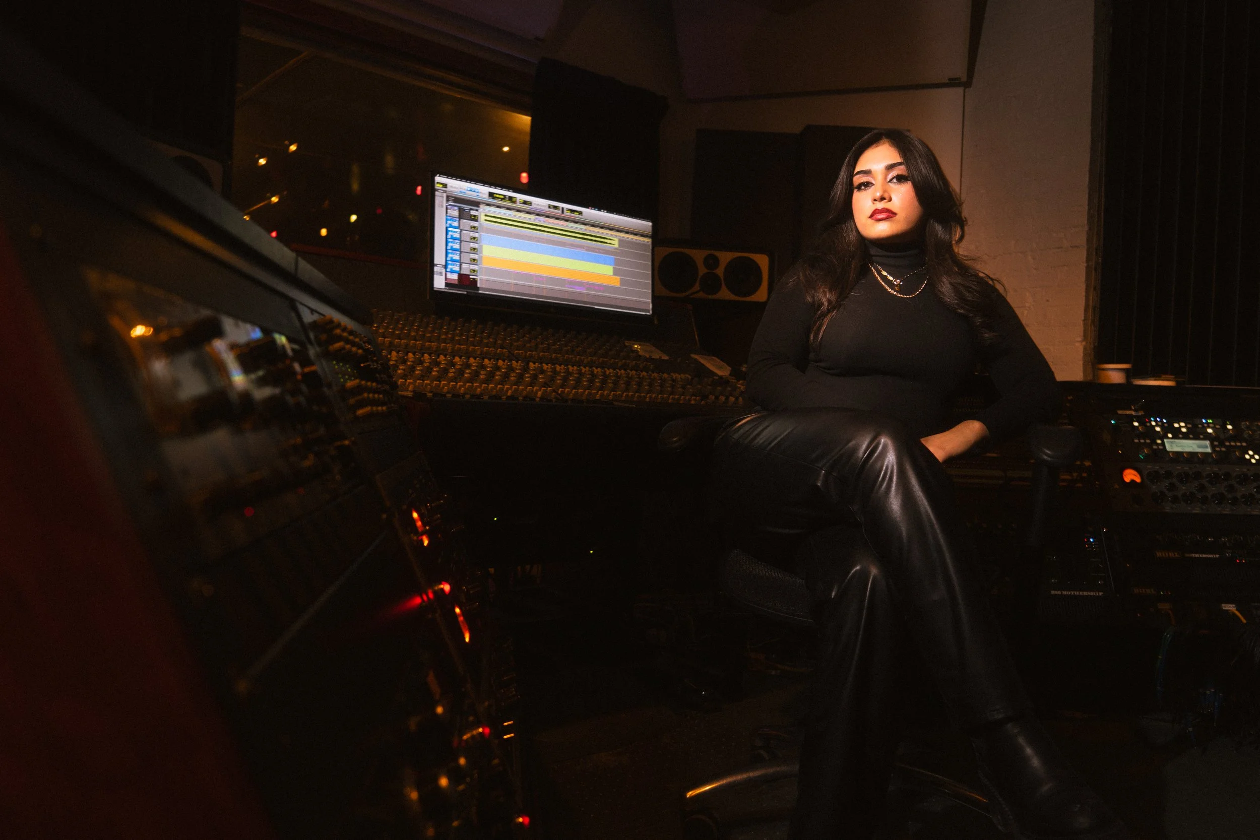 A woman sitting in a music recording studio near professional audio equipment and a computer monitor displaying digital audio work station software.