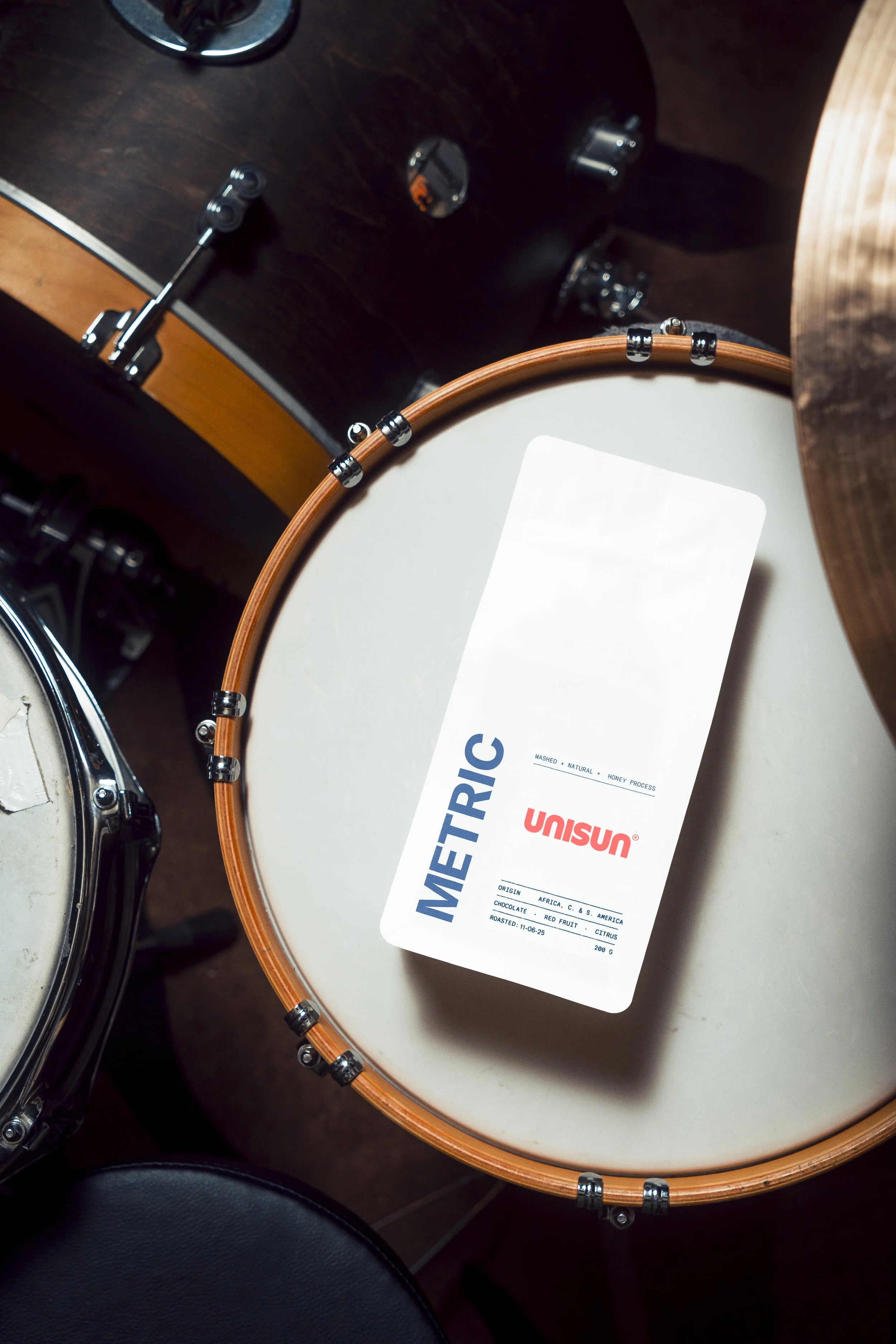Product photo of white drum with a label indicating it contains honey process coffee beans, and a part of a percussion instrument with a wooden rim and metal screws.