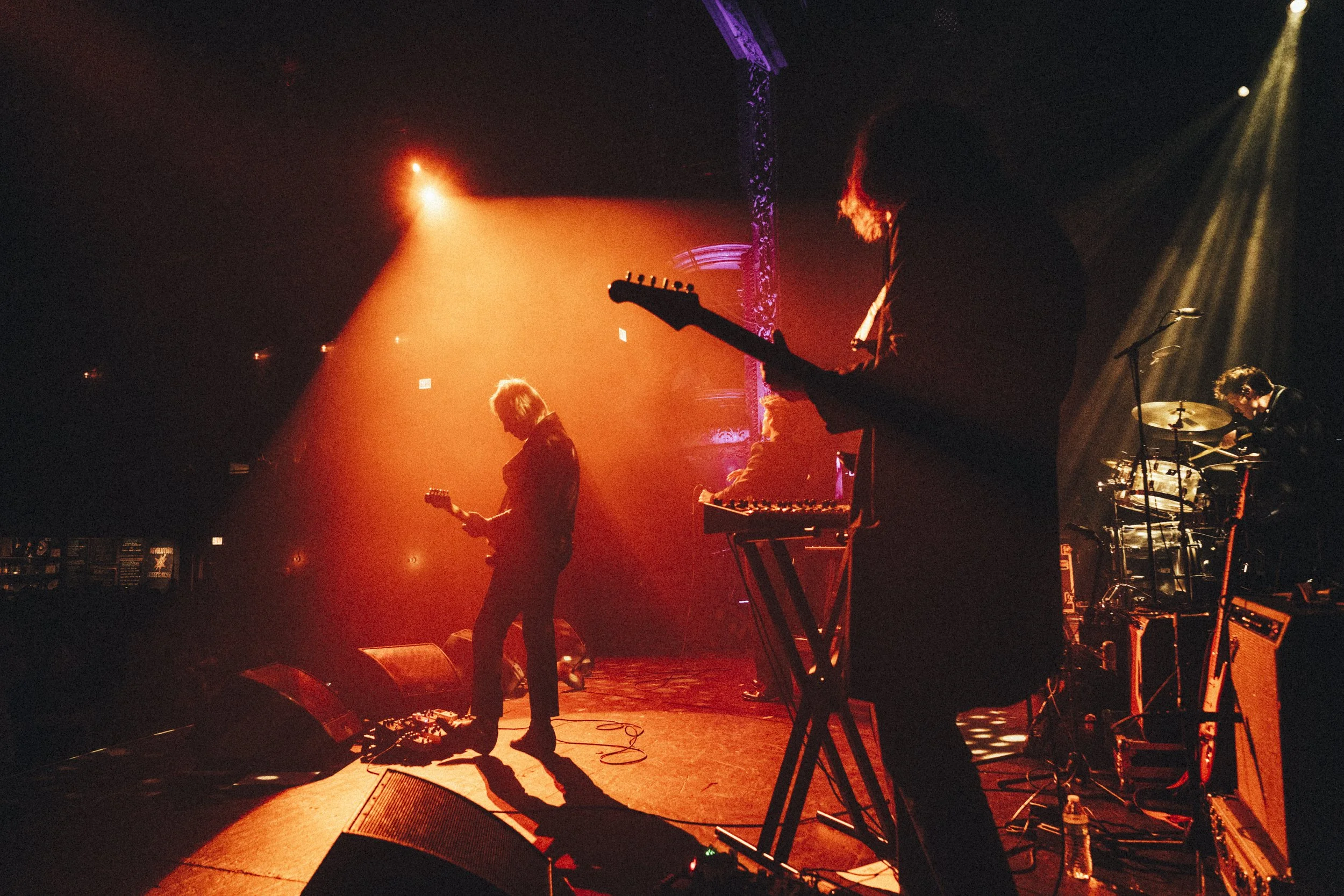 A band performing on stage with orange and purple lighting, featuring a guitarist, keyboardist, and drummer.