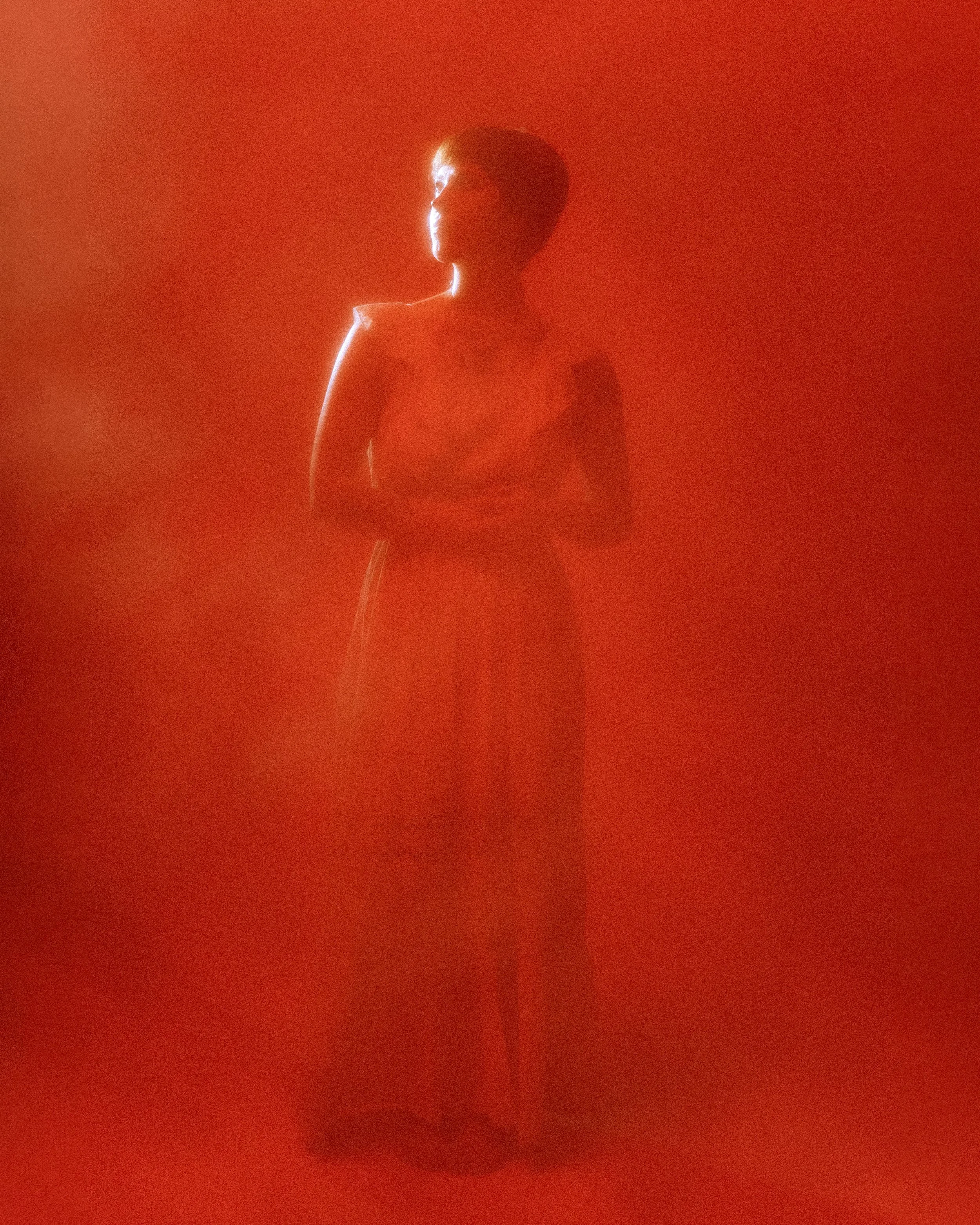 An atmospheric and dramatic portrait of a person with short hair stands against a red background, illuminated by side lighting, creating a silhouette effect.