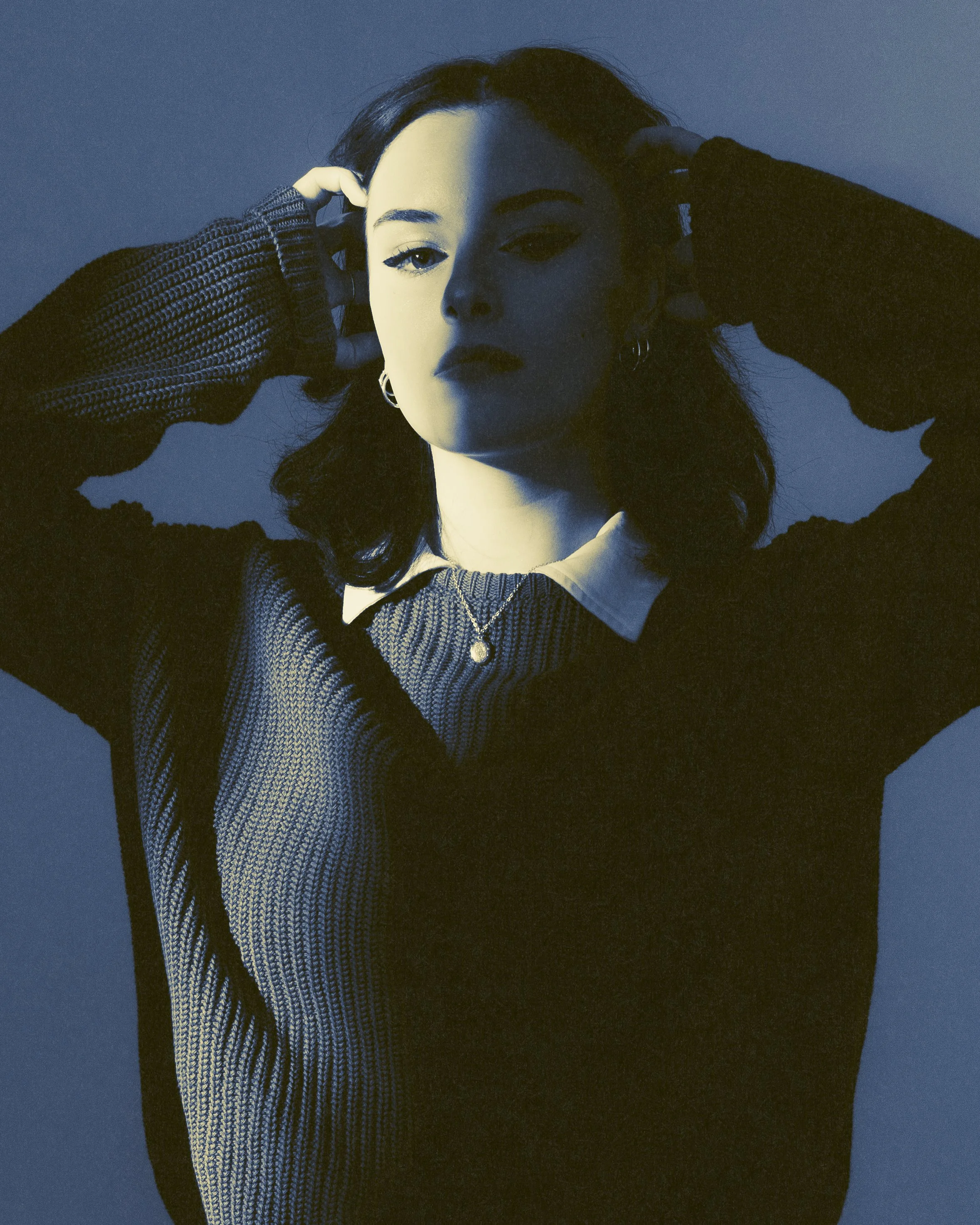A woman with dark hair posing against a blue background, with half her face in shadow, wearing a dark sweater, a white collared shirt, and layered necklaces.