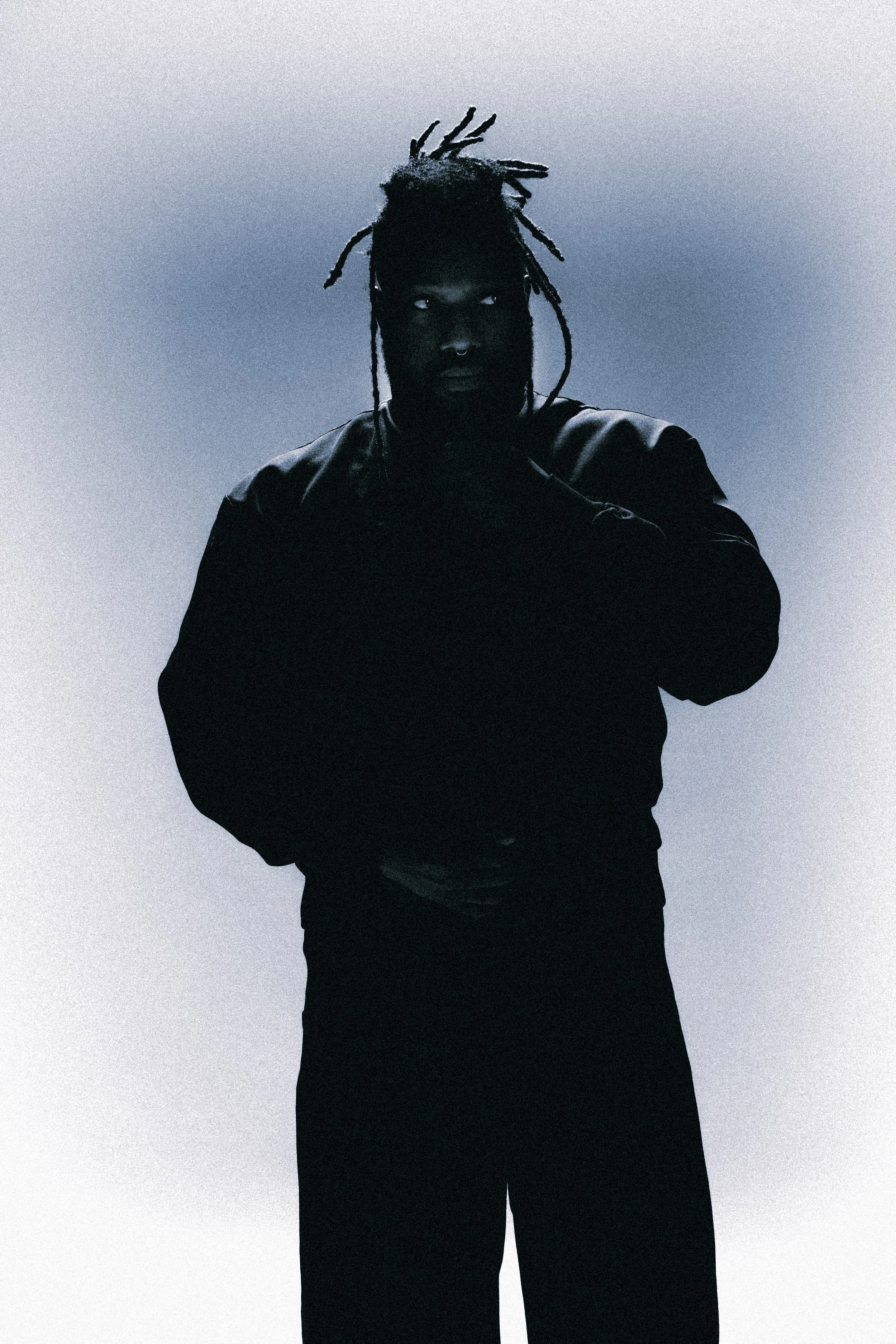 Person with dreadlocks standing in front of a plain background, wearing a dark jacket best for music promotion or tour poster or eye catching social media post