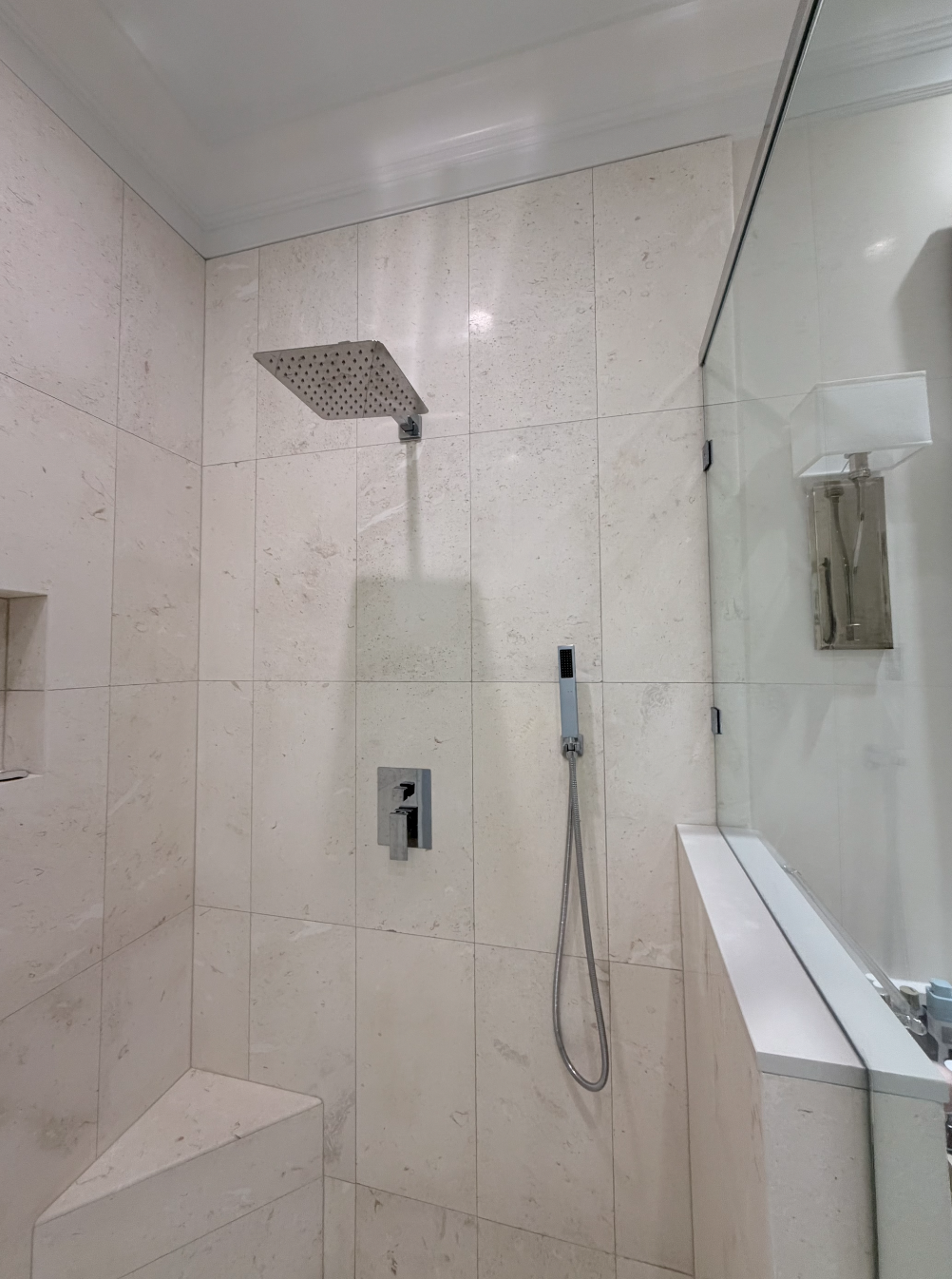 Modern shower in a bathroom with beige tiled walls, featuring a square rain showerhead mounted on the ceiling, a handheld shower with a hose on the wall, a built-in niche in the corner, and a glass shower door.