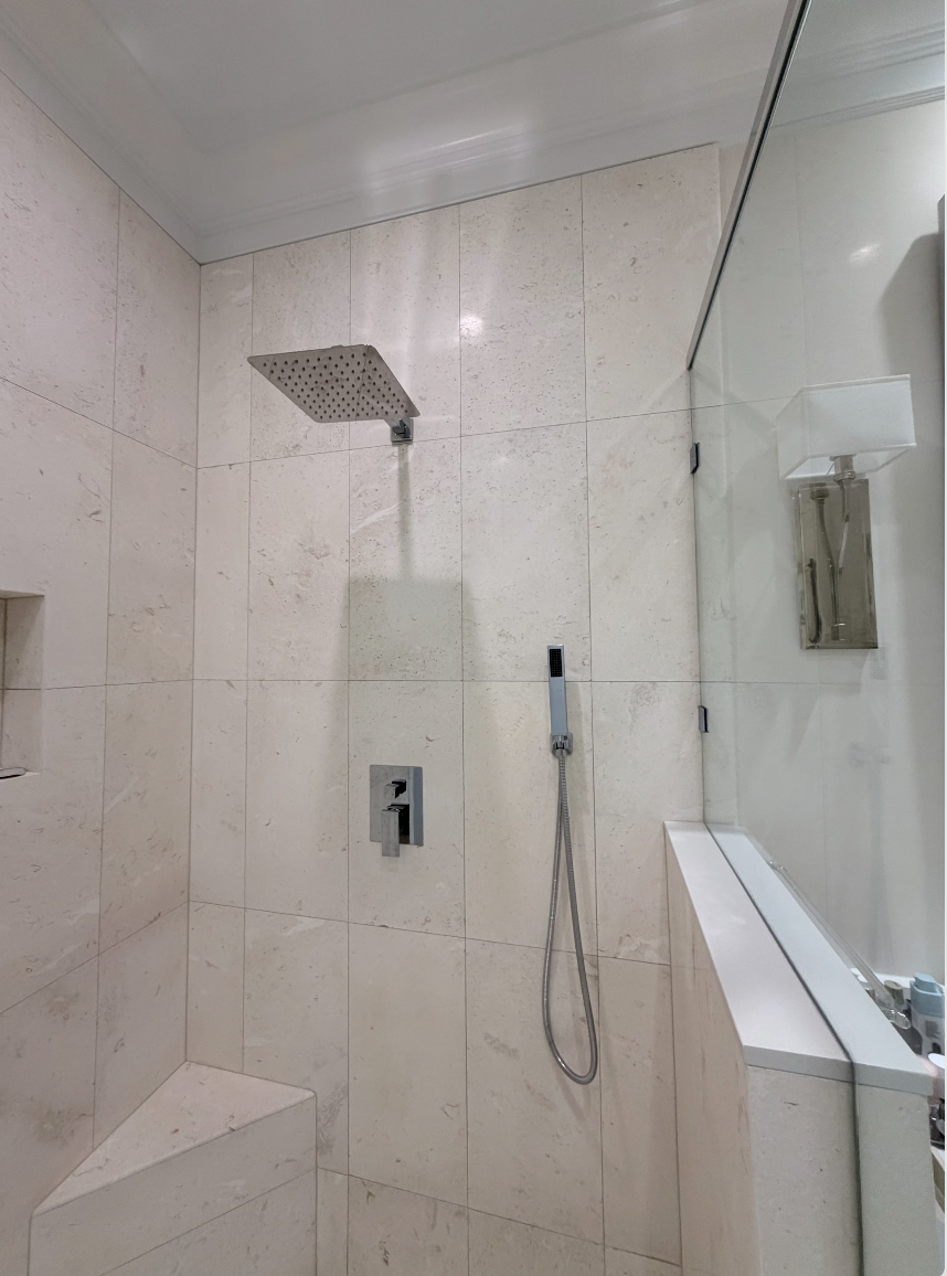 A modern shower with beige tiled walls, a square rainfall showerhead, a handheld shower and a control panel, with a glass partition and a white ledge.