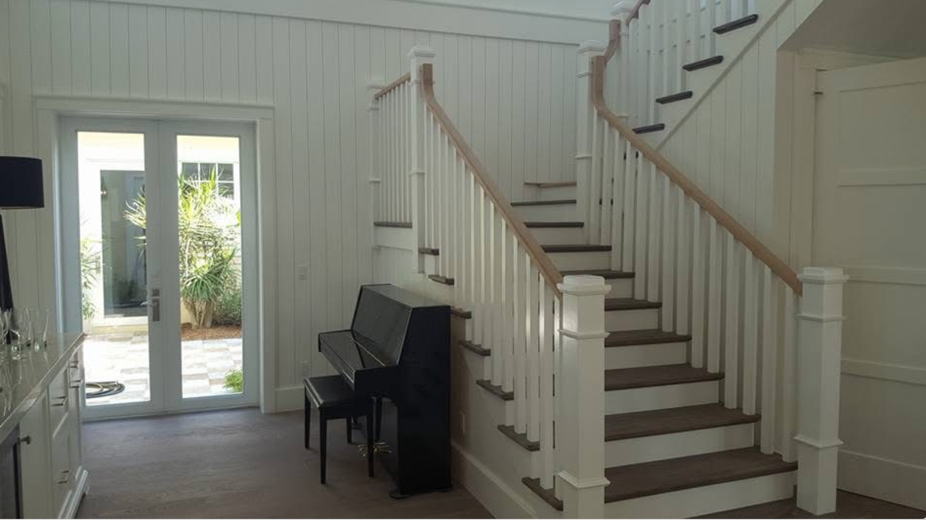 Interior view of a house with a staircase, a black upright piano, and a glass door leading outside to a patio with plants.